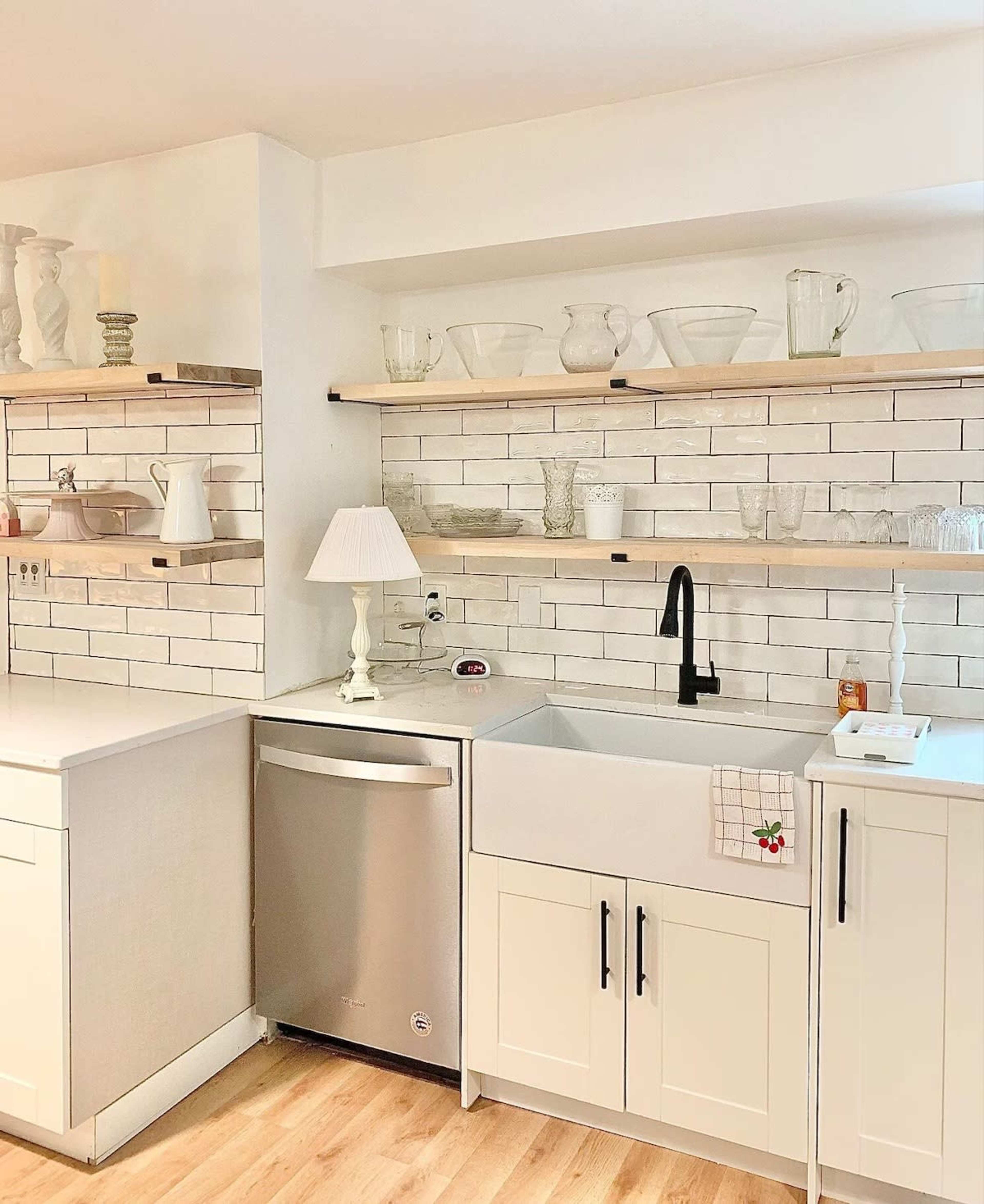A modern kitchen features a farmhouse sink, black faucet, open shelves displaying glassware, and light-colored cabinetry.