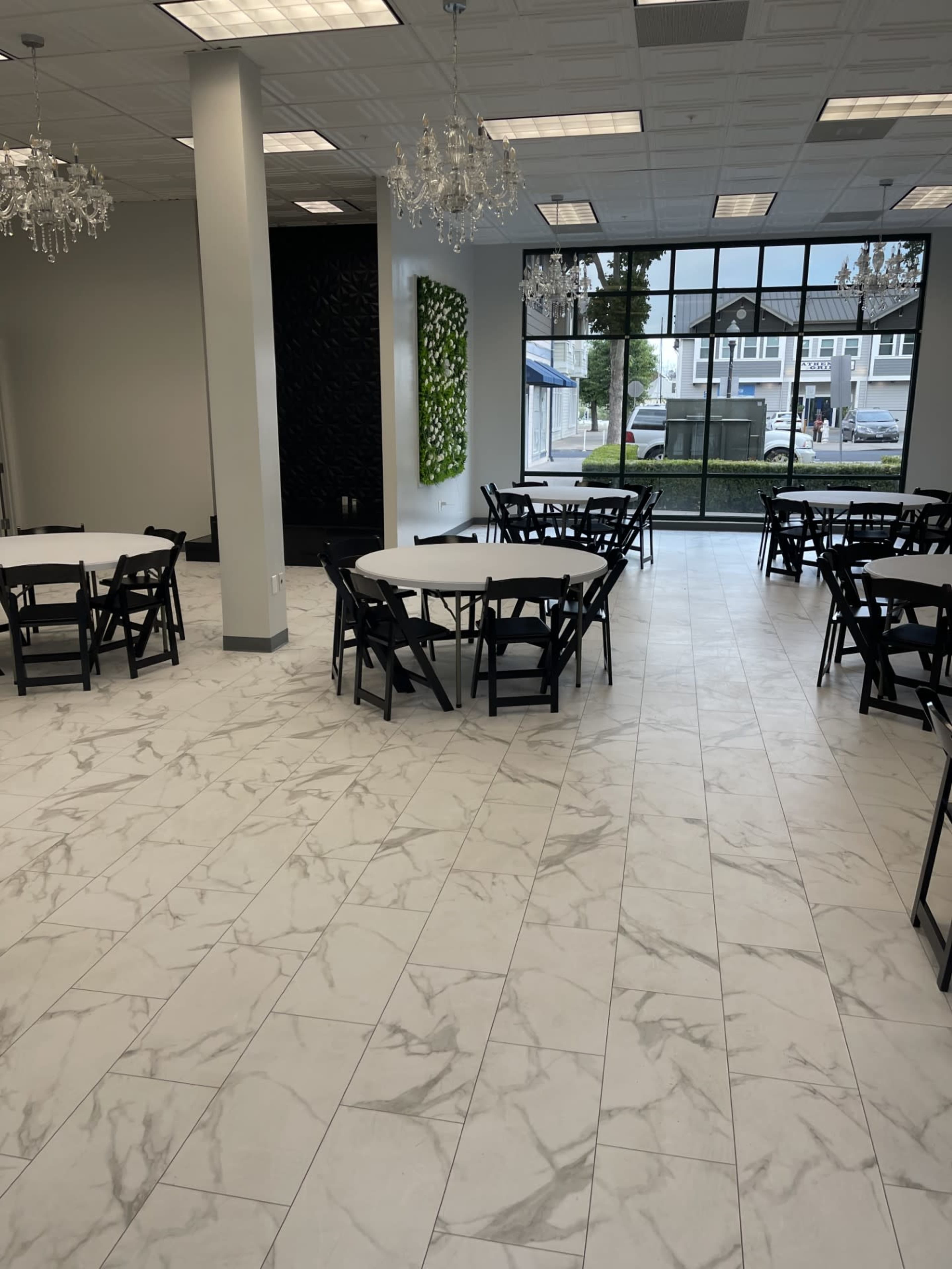 A spacious room with round tables and black folding chairs arranged on a marble-patterned tile floor, featuring large windows that let in natural light.