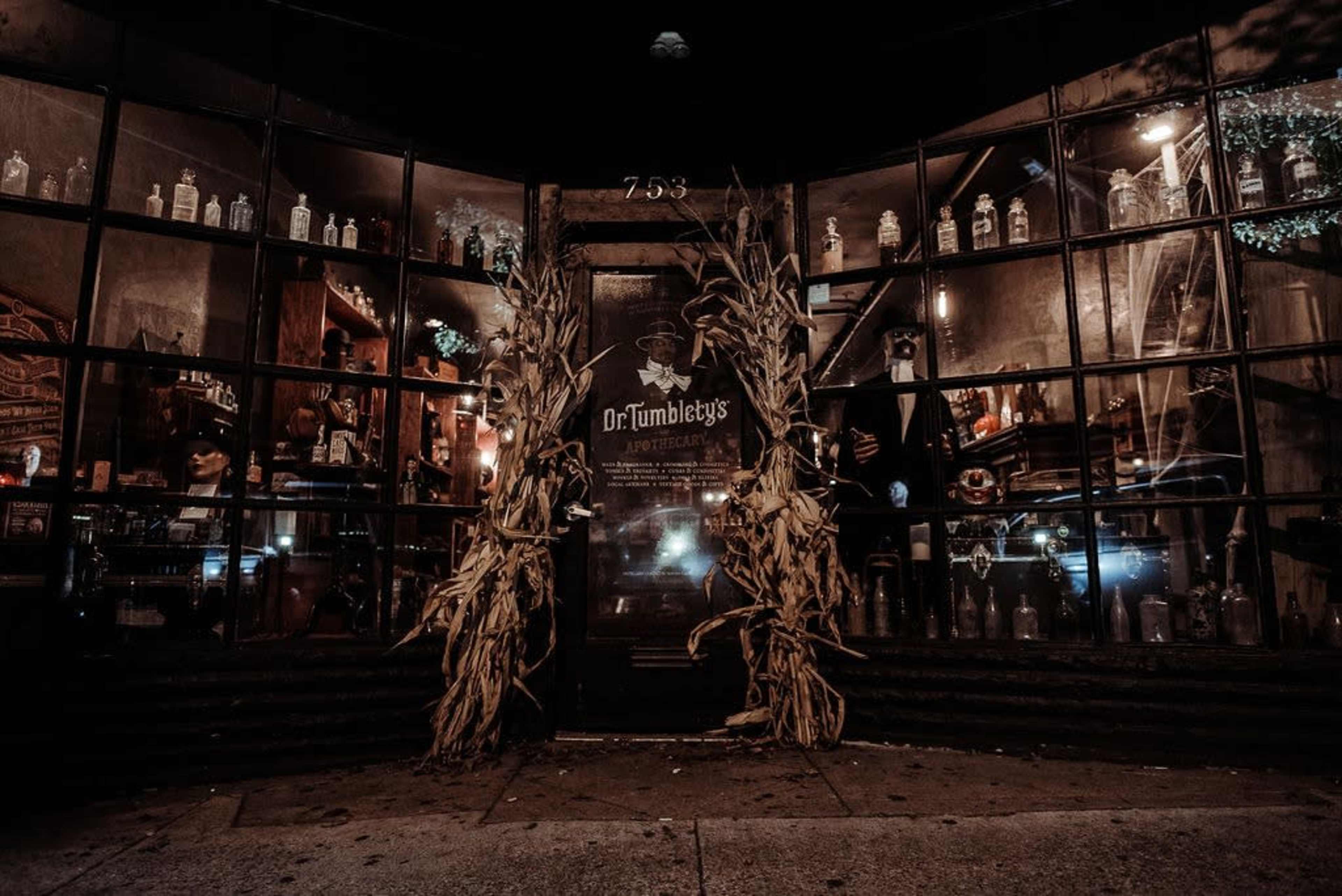 The entrance of Dr. Tumbleby's is adorned with dried corn stalks and features large glass windows showcasing various bottles and decorative items inside.