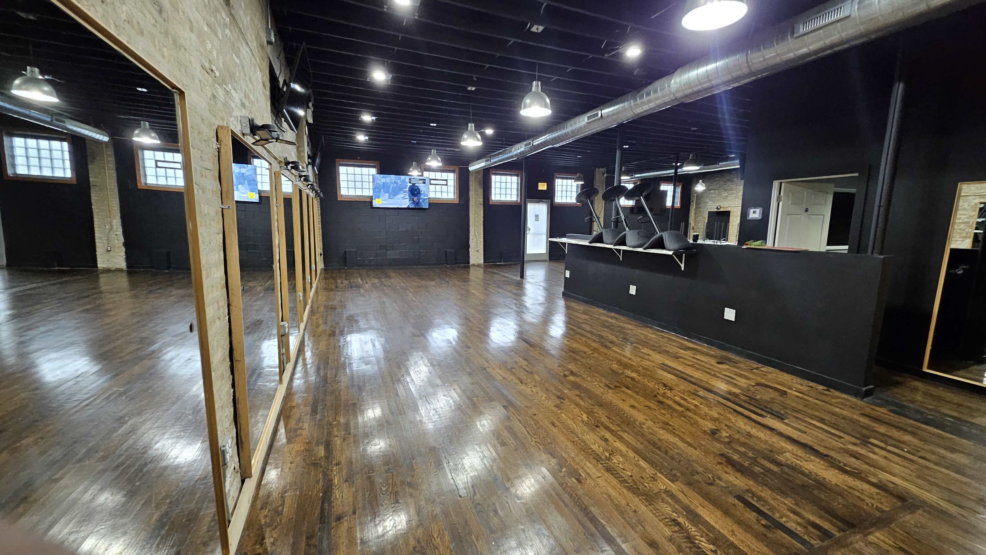 The image shows an empty fitness studio with wooden floors, large mirrors, and a black wall featuring exercise equipment.