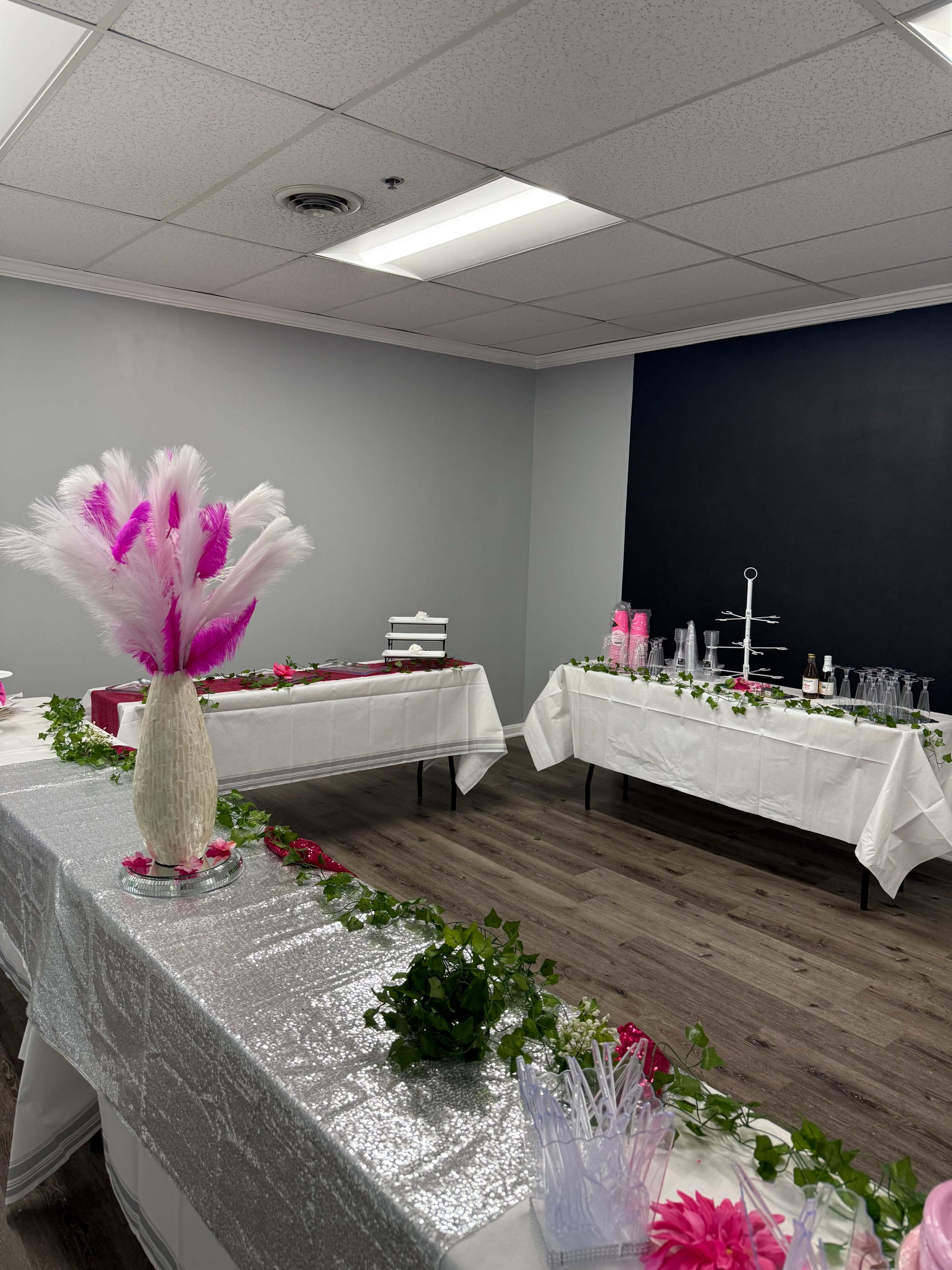 The image shows a decorated room with tables draped in white cloth, featuring pink and white floral arrangements, greenery, and event supplies set up for a celebration.