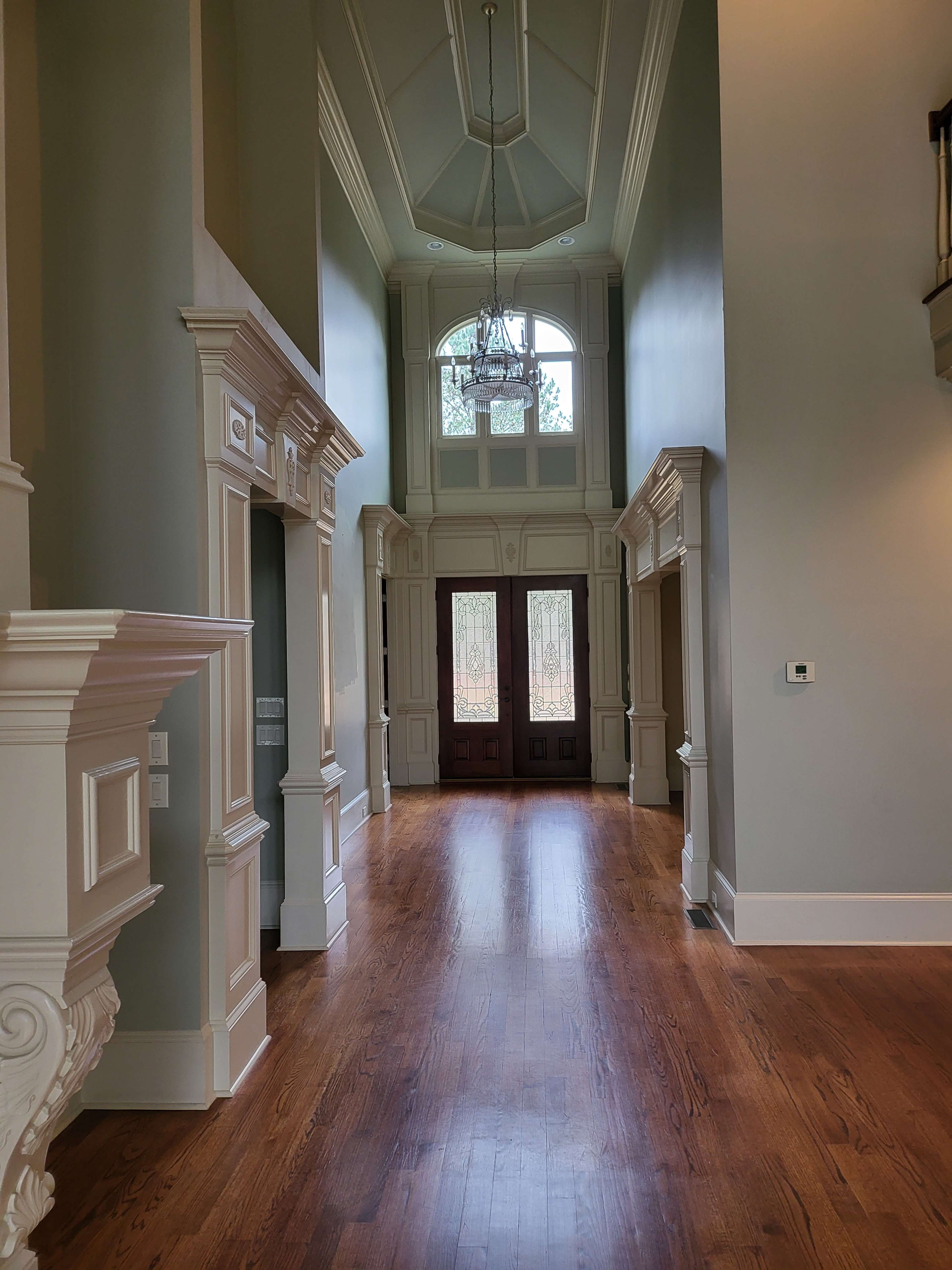 A brightly lit hallway in a home, featuring tall ceilings, ornate moldings, and large double doors at the end leading to an outdoor view.