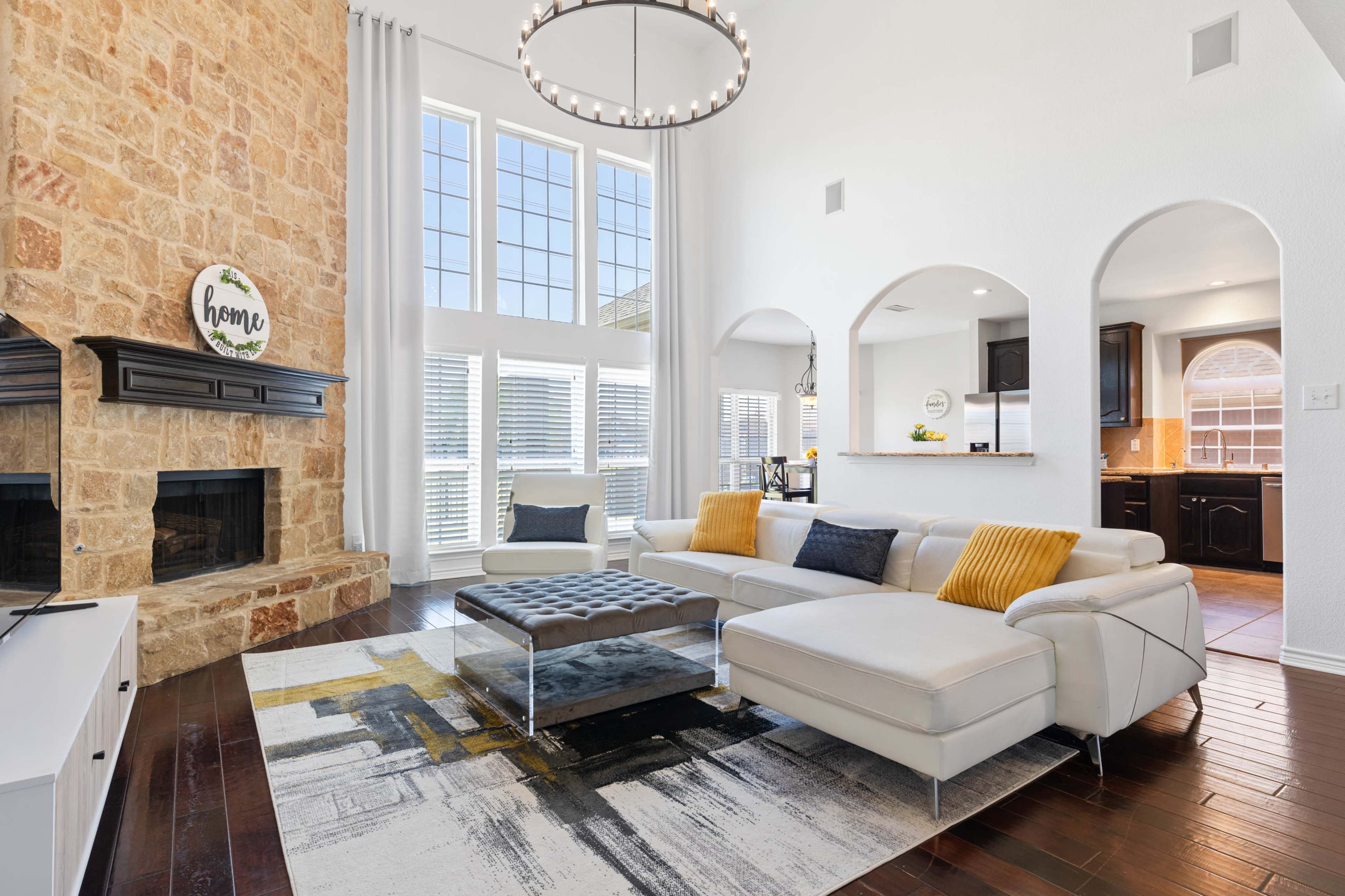 A modern living room with a stone fireplace, large windows, and a light-colored sectional sofa positioned on a patterned area rug.