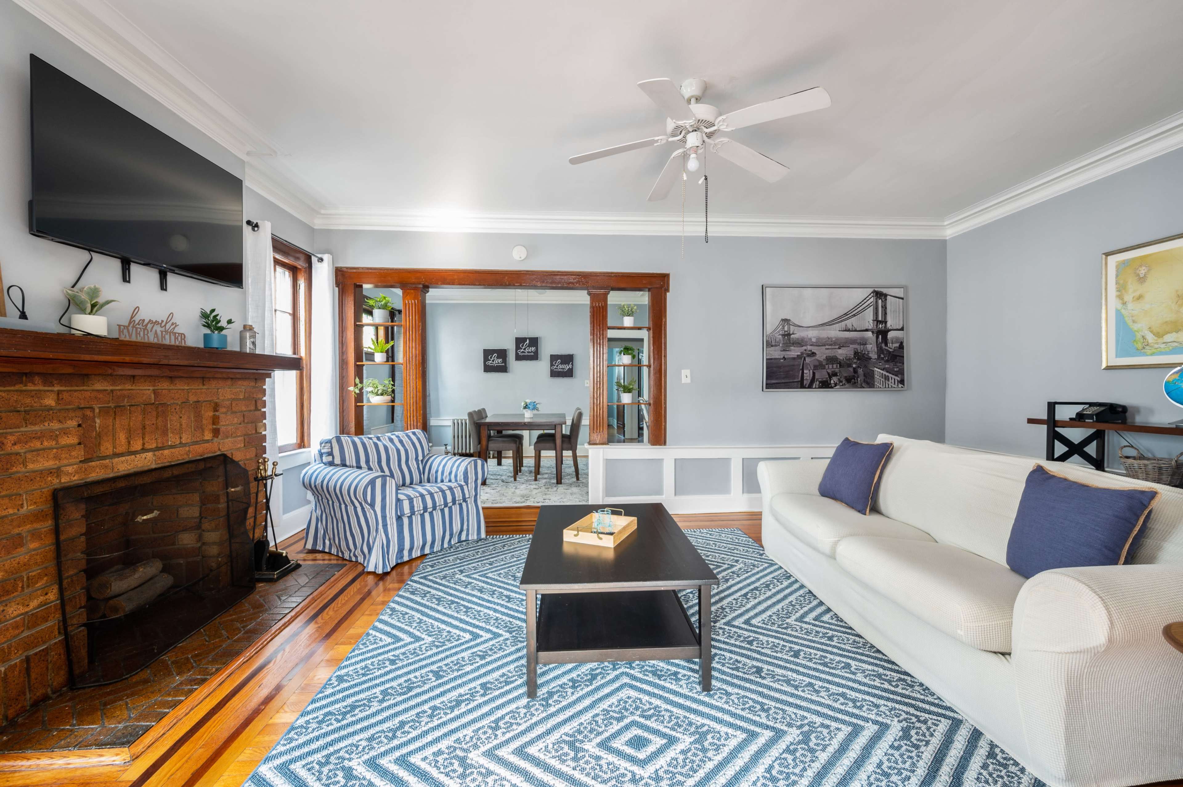 The living room features a white sofa, a striped blue chair, a coffee table, and a fireplace, with a television mounted on the wall and art displayed around the room.
