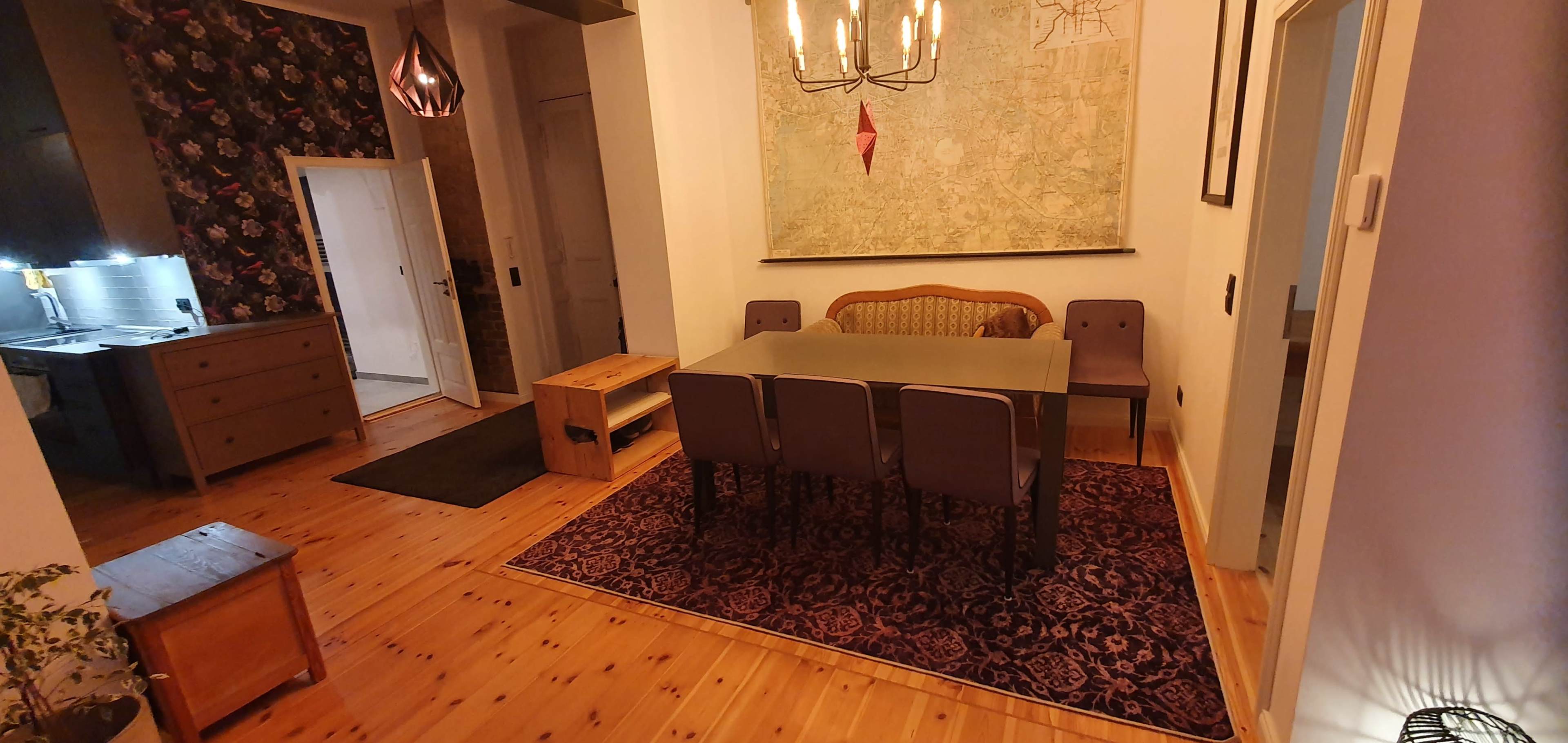 The image shows a dining area featuring a table with four chairs, a light fixture above, and a large map hanging on the wall, situated on a patterned rug.
