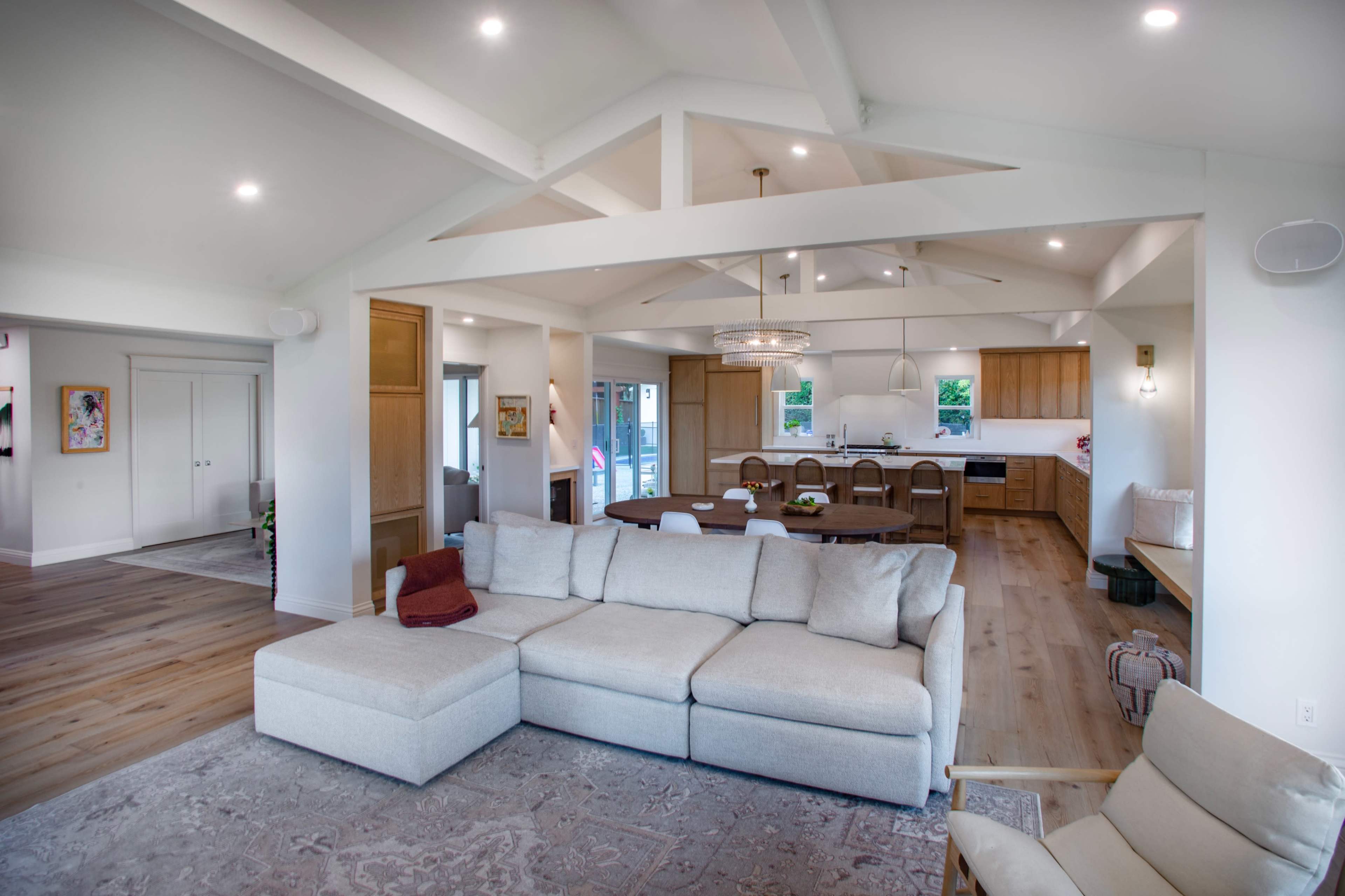 The image shows a modern living space featuring a light-filled room with a large sectional sofa, a dining area with a wooden table, and a kitchen in the background.