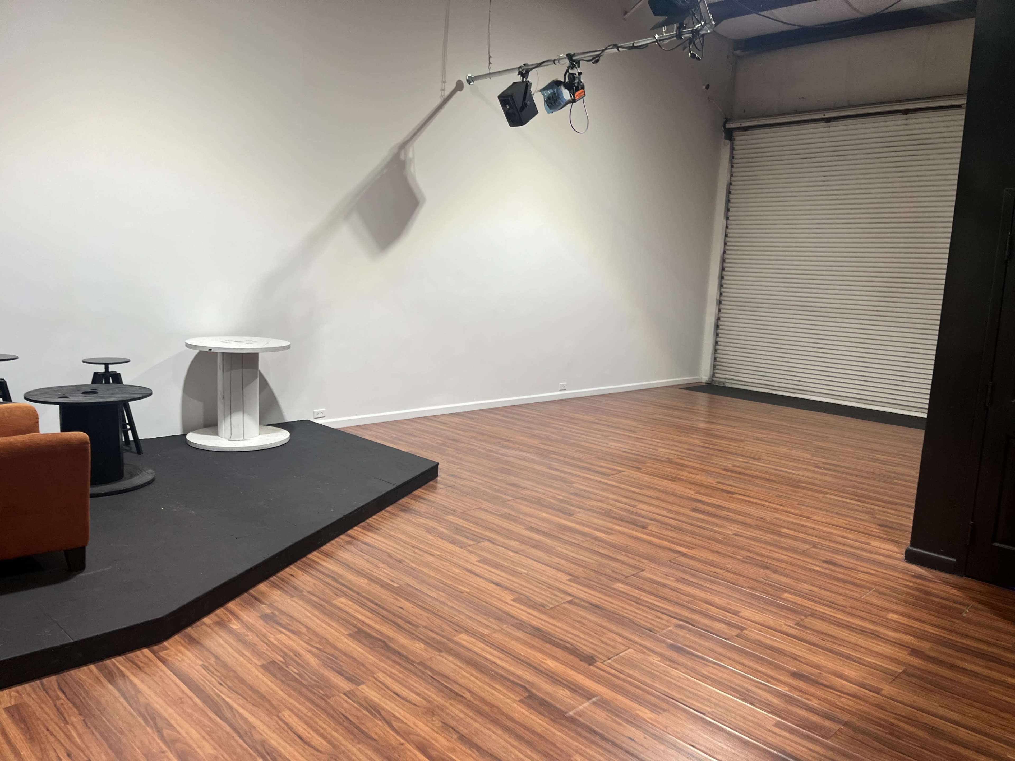 A spacious, empty room with wooden flooring, a small stage area, and a rolling door in the background.