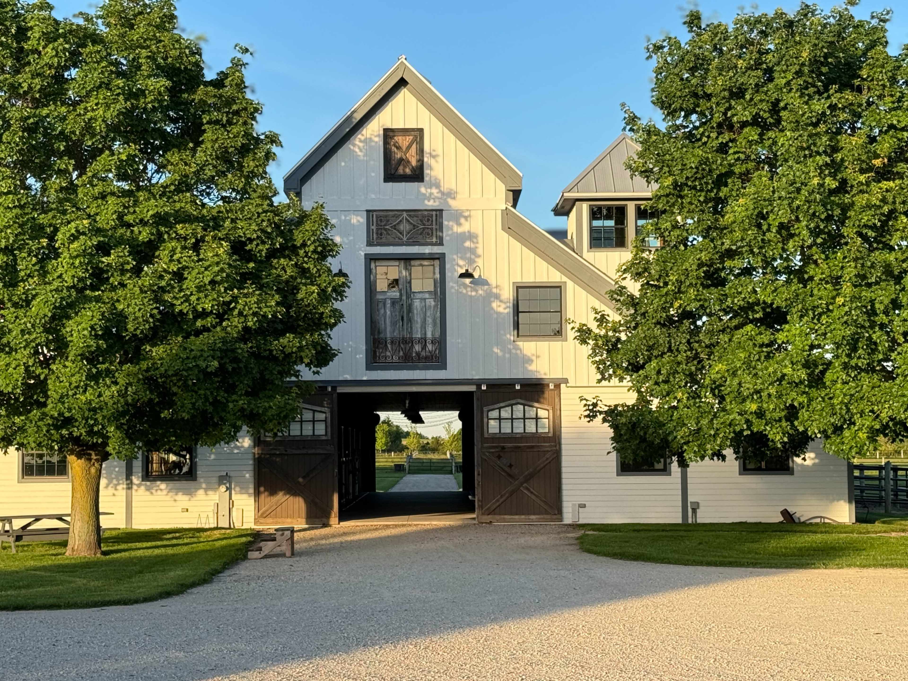 A white barn with large wooden doors is flanked by two trees, set against a clear blue sky.