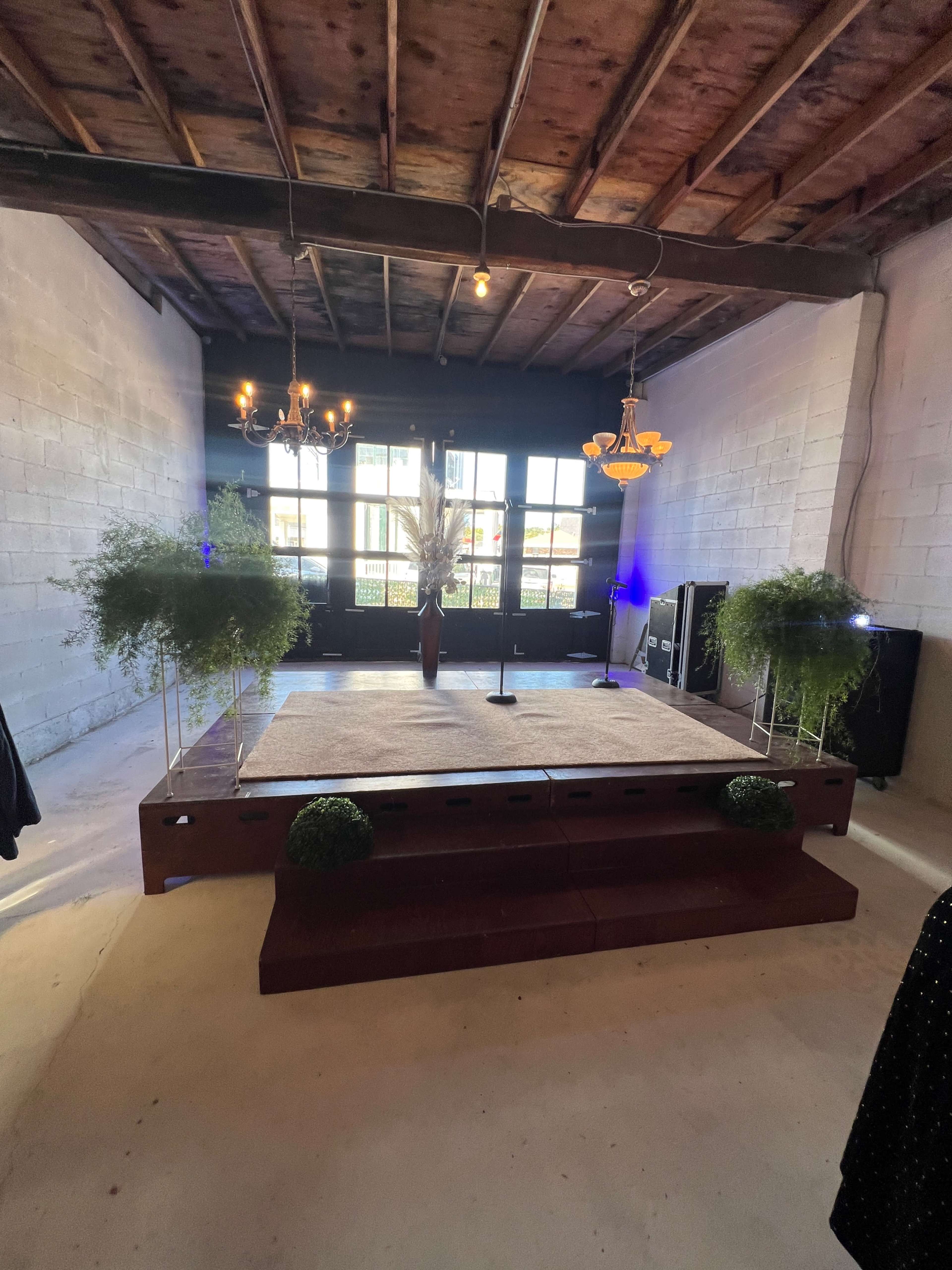 The image shows a simple stage setup in a room with exposed wooden beams, decorated with potted plants and lighting fixtures.