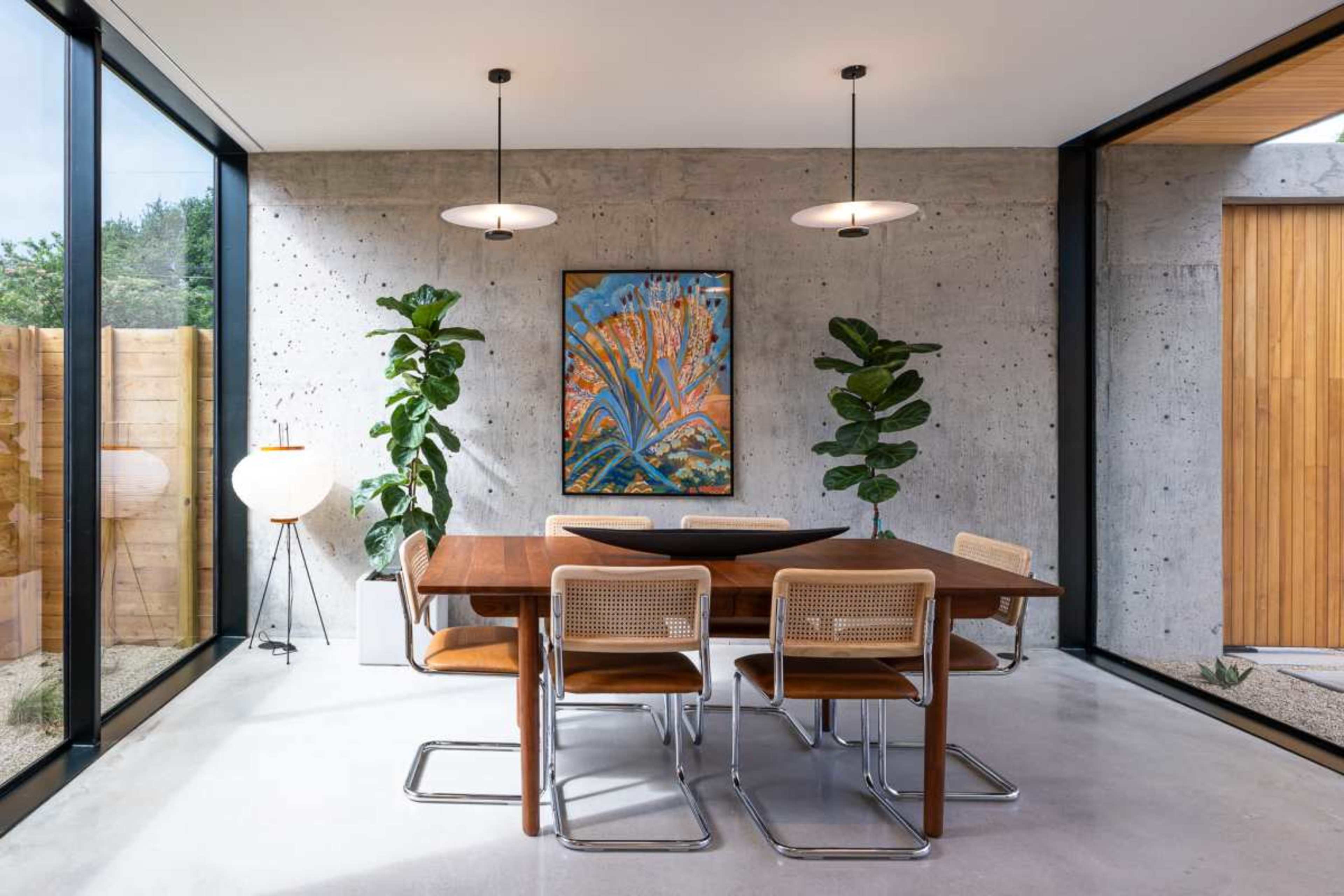 A modern dining area features a wooden table with four chairs, surrounded by large windows, plants, and a colorful artwork on the concrete wall.