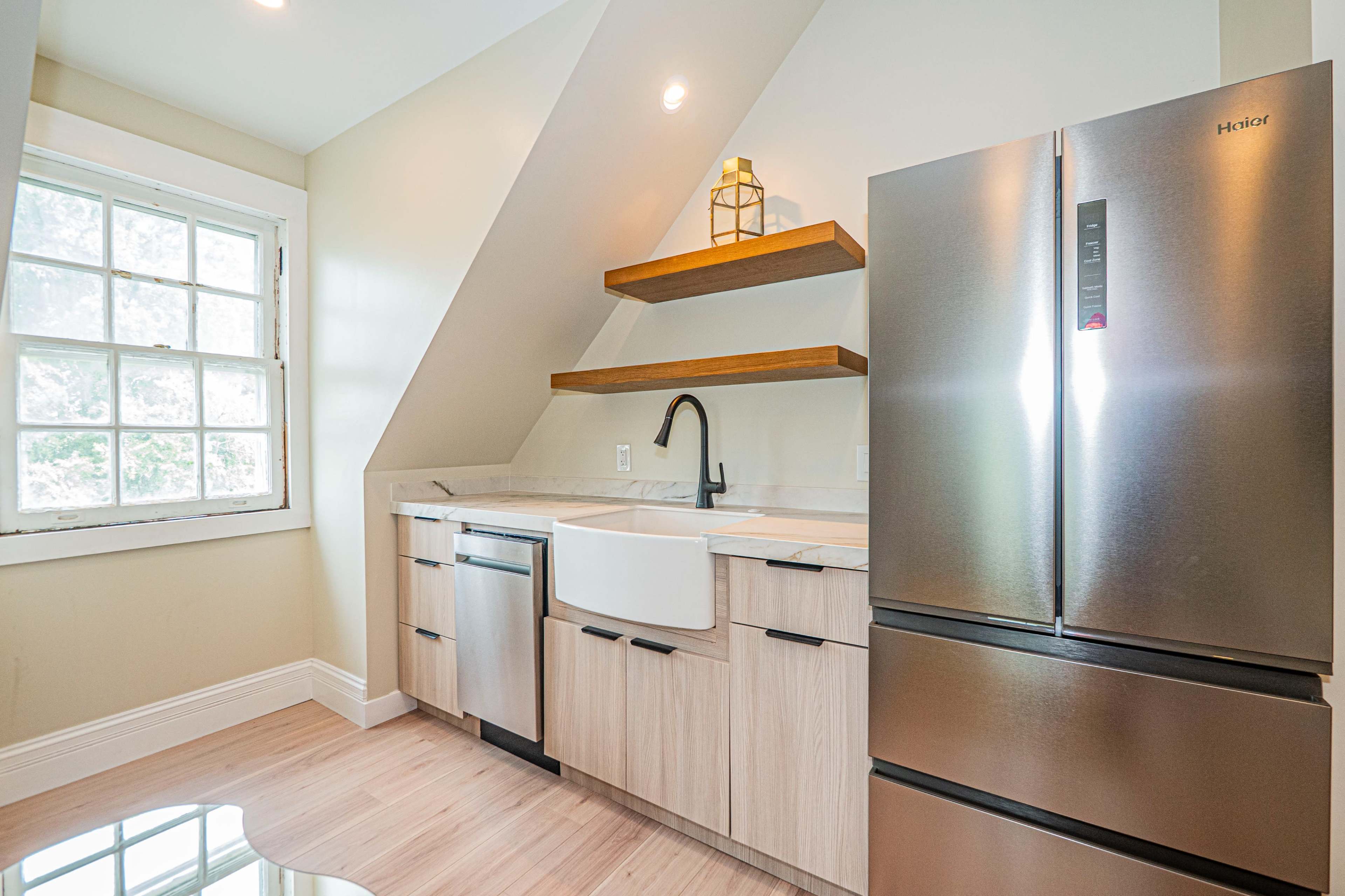 The image shows a modern kitchen with an angled ceiling, featuring a stainless steel refrigerator, a farmhouse sink, and wooden shelves above a light-colored countertop.