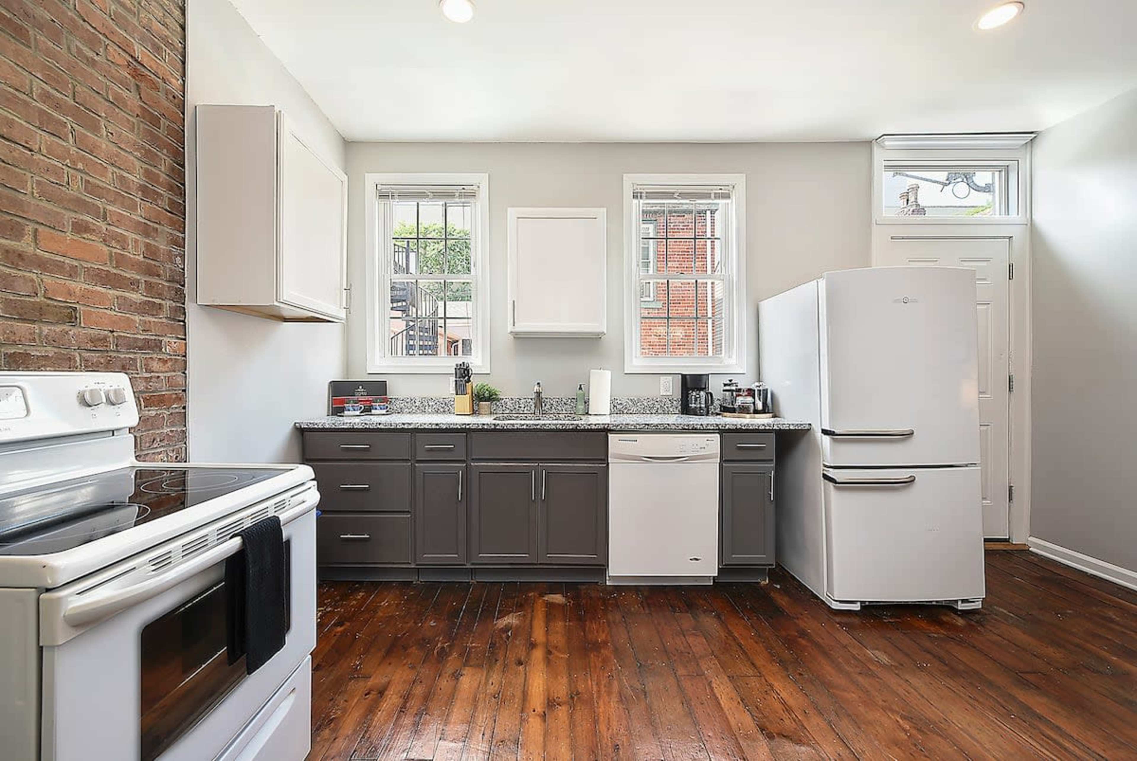The kitchen features a combination of brick and painted walls, with a stove, refrigerator, and dishwasher arranged along a countertop.