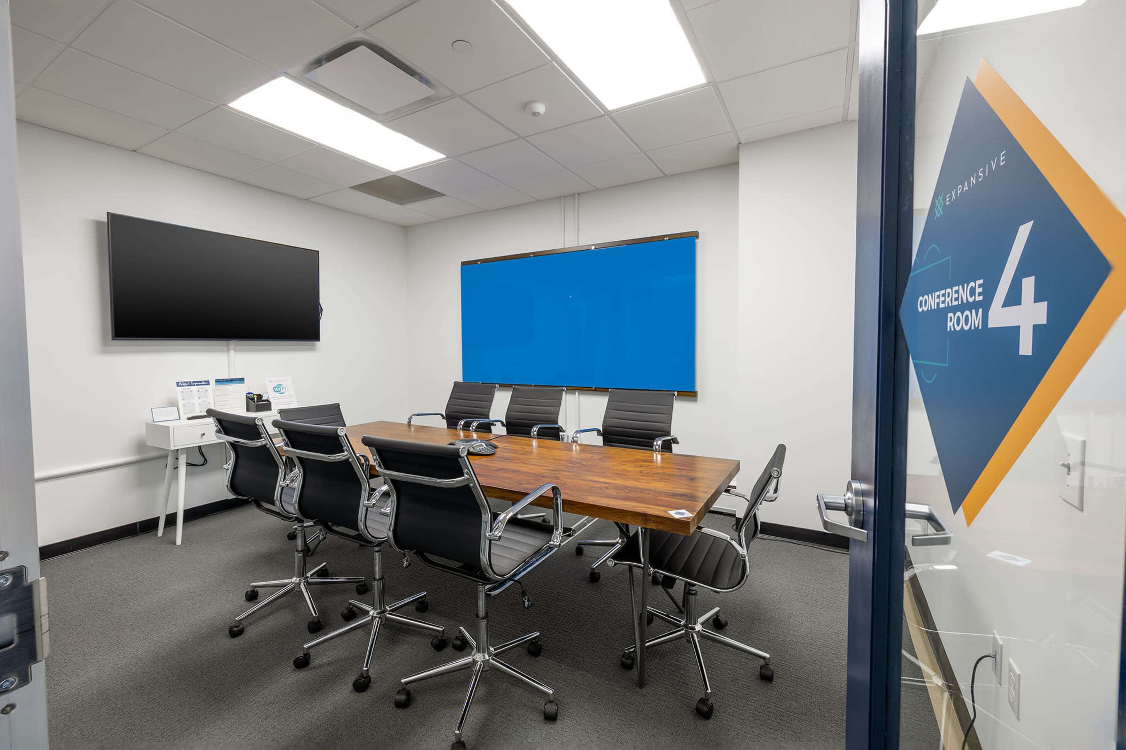 The image shows a modern conference room with a wooden table, black chairs, and two large screens on the walls.