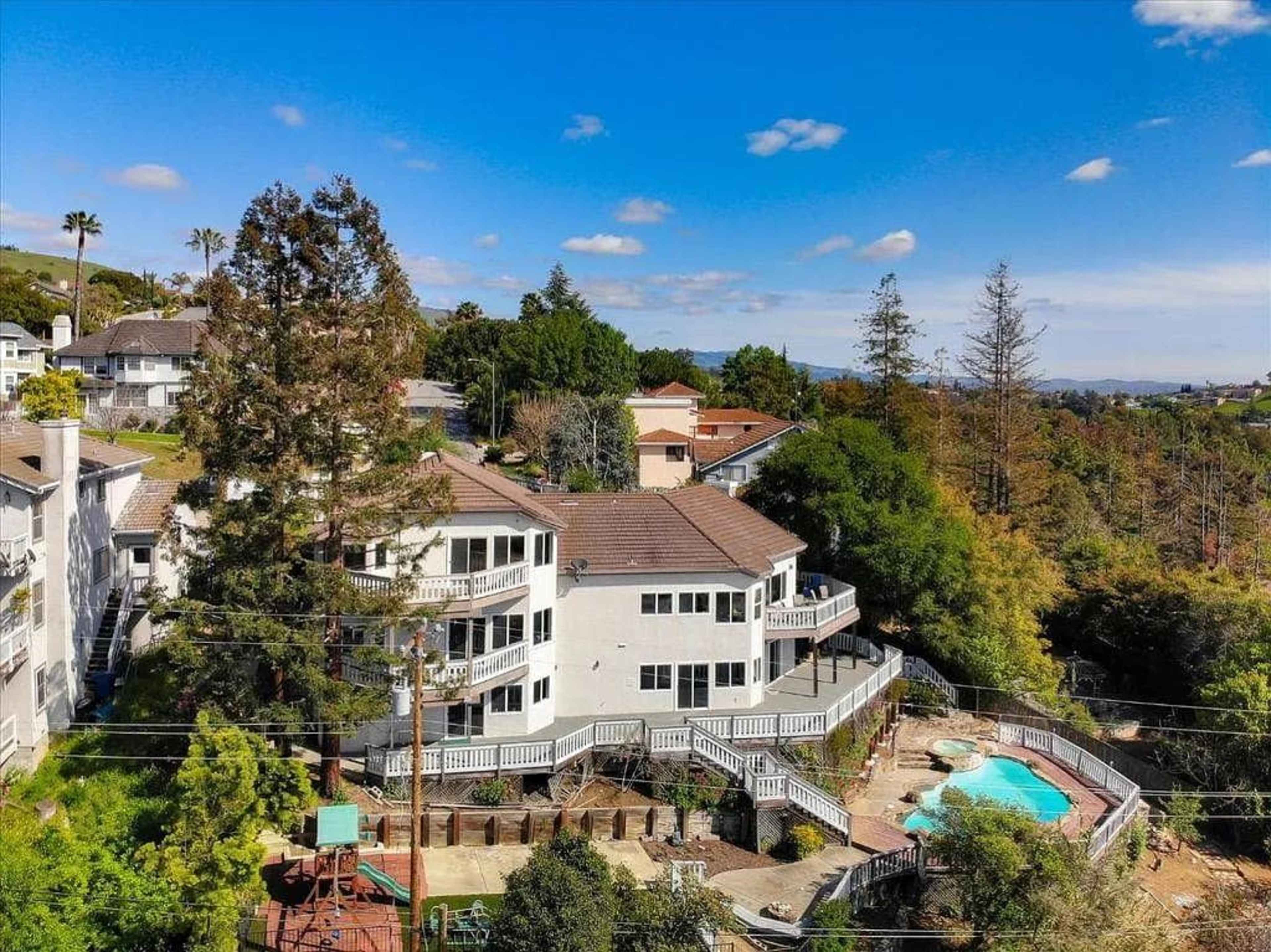 A large white house with multiple balconies and a swimming pool is surrounded by trees and other residential buildings.