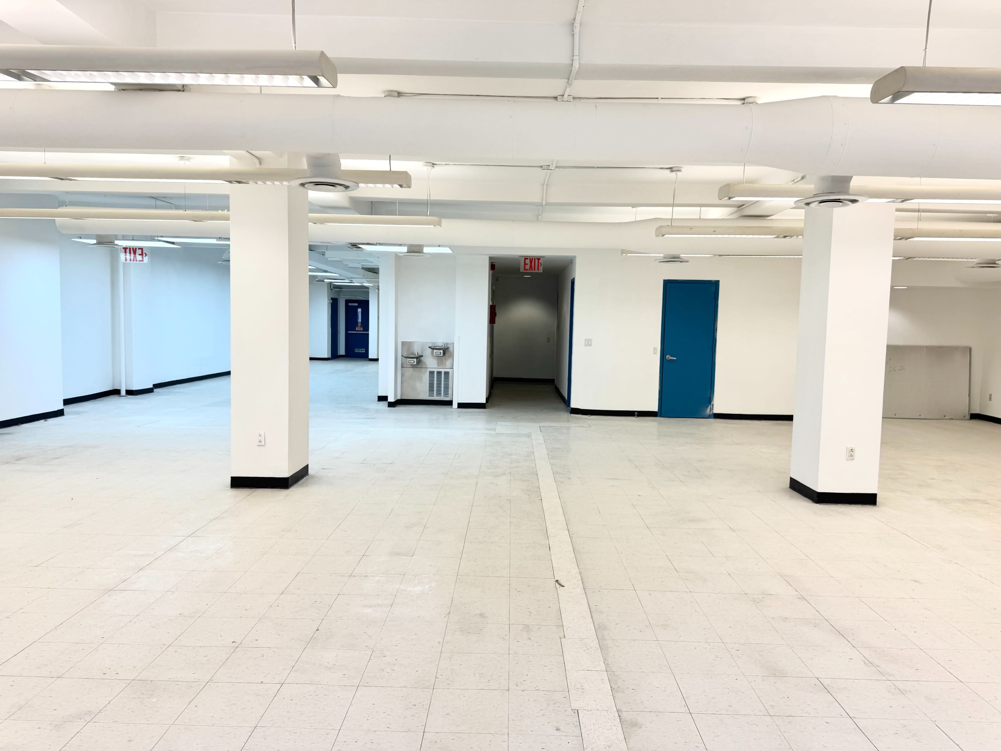 The image shows a spacious, empty room with white walls, tiled flooring, and several columns supporting the ceiling.
