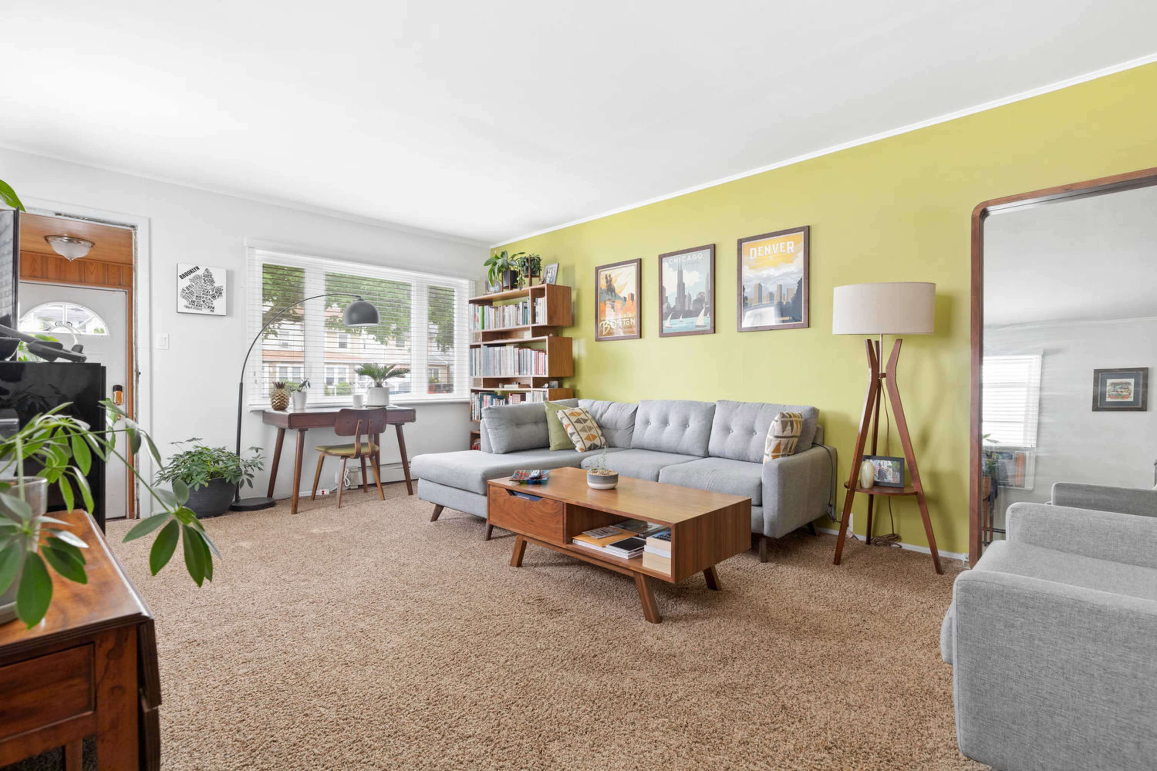 A cozy living room features a gray sofa, a wooden coffee table, and a small workspace by a window, with green walls and various decorative plants.