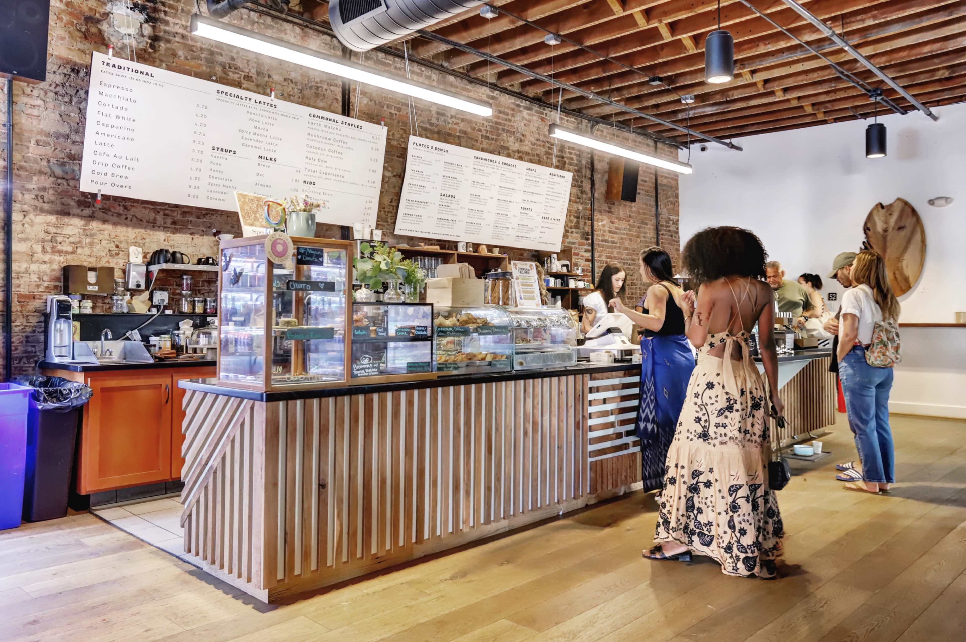 A bustling café features a wooden counter with a display of pastries, and customers are lined up while baristas prepare drinks behind the counter.