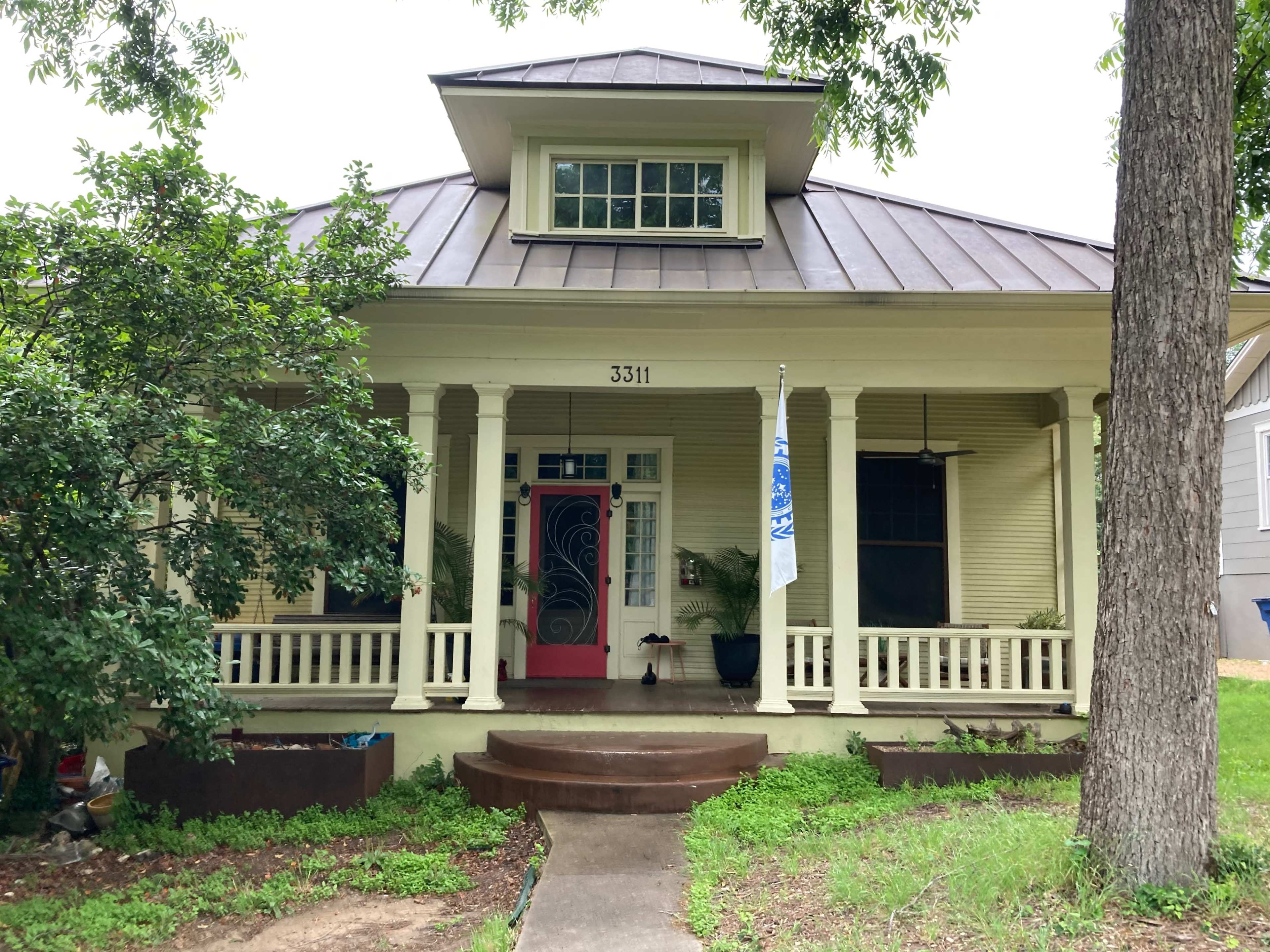 A two-story house with a front porch, featuring a red door and a metal roof, surrounded by greenery.