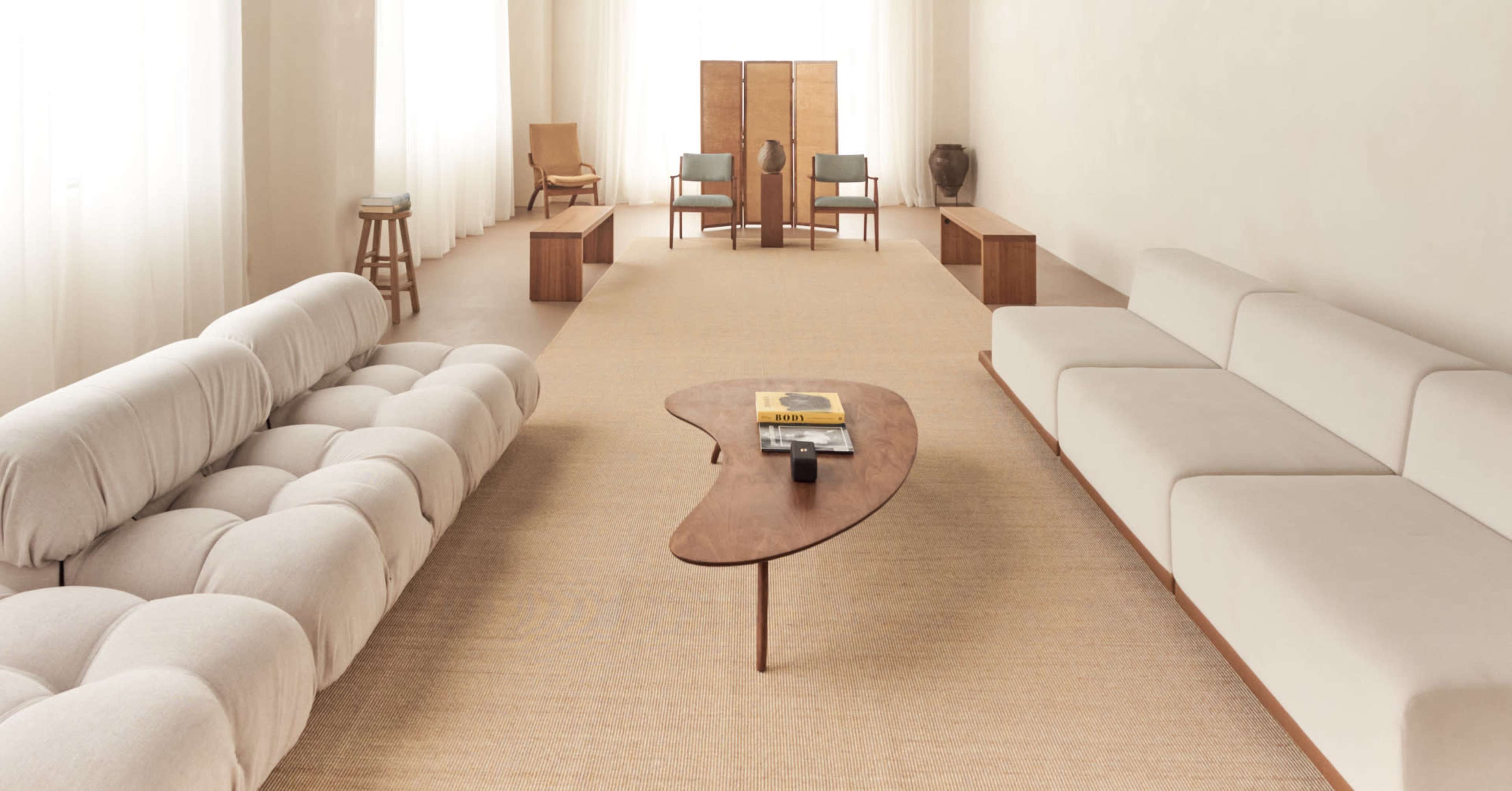 The image shows a spacious, minimalist living room featuring a light-colored sofa, a sculptural wooden coffee table, and two chairs arranged around a central wooden screen.