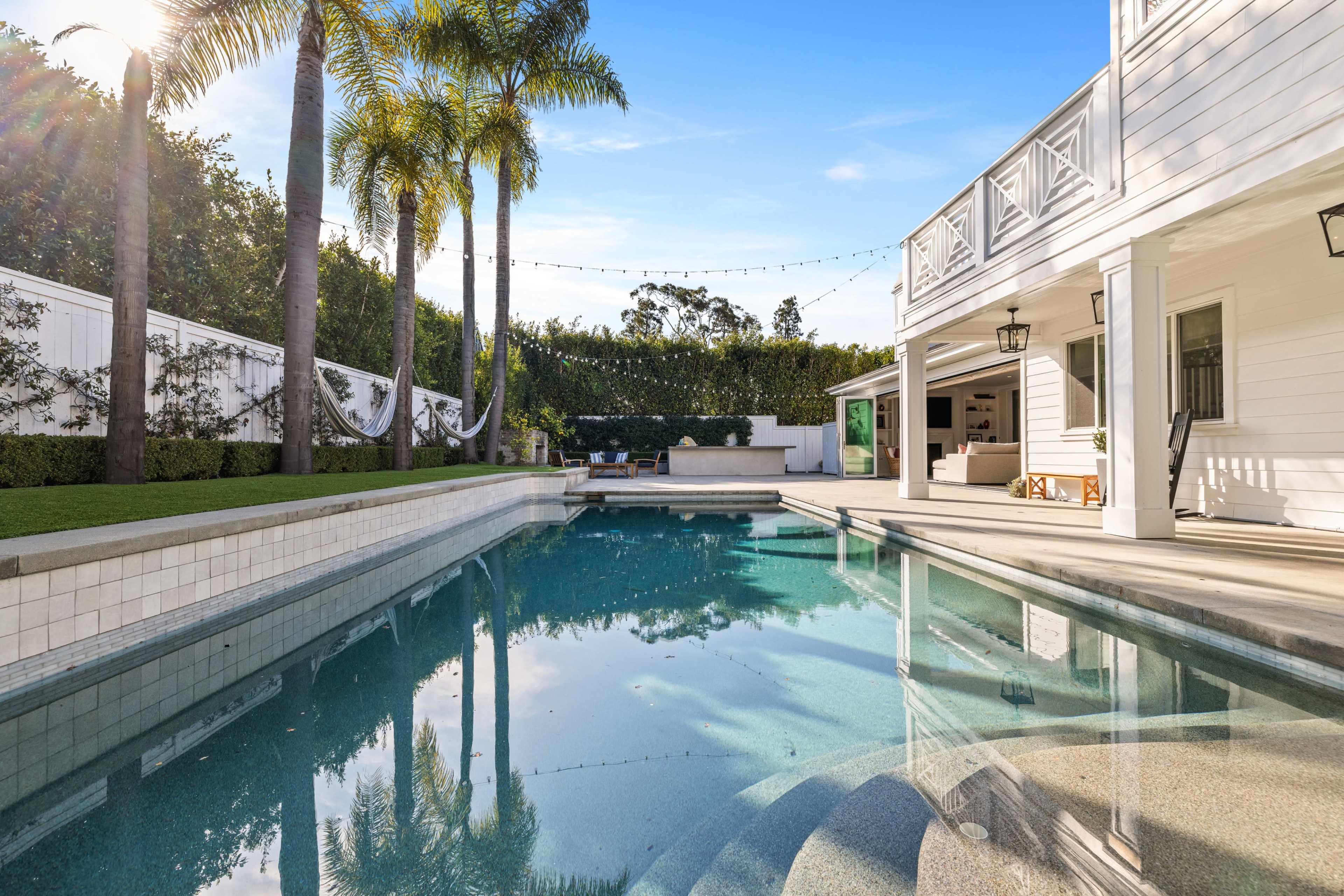 The image shows a clear swimming pool surrounded by tropical plants and a modern house with outdoor seating areas.