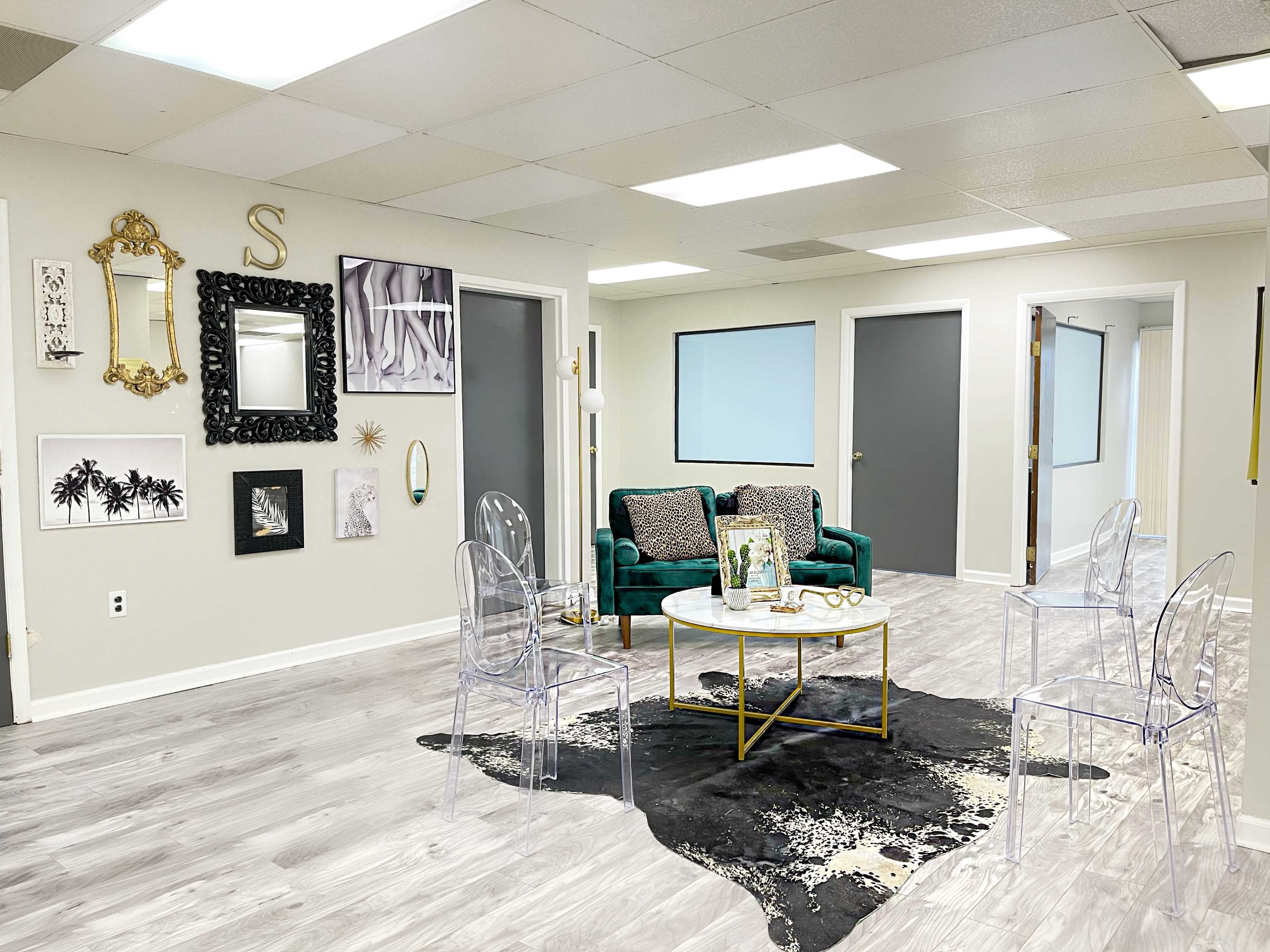 A spacious, well-lit room featuring a green velvet sofa, a round coffee table, several transparent chairs, and an array of framed wall art.
