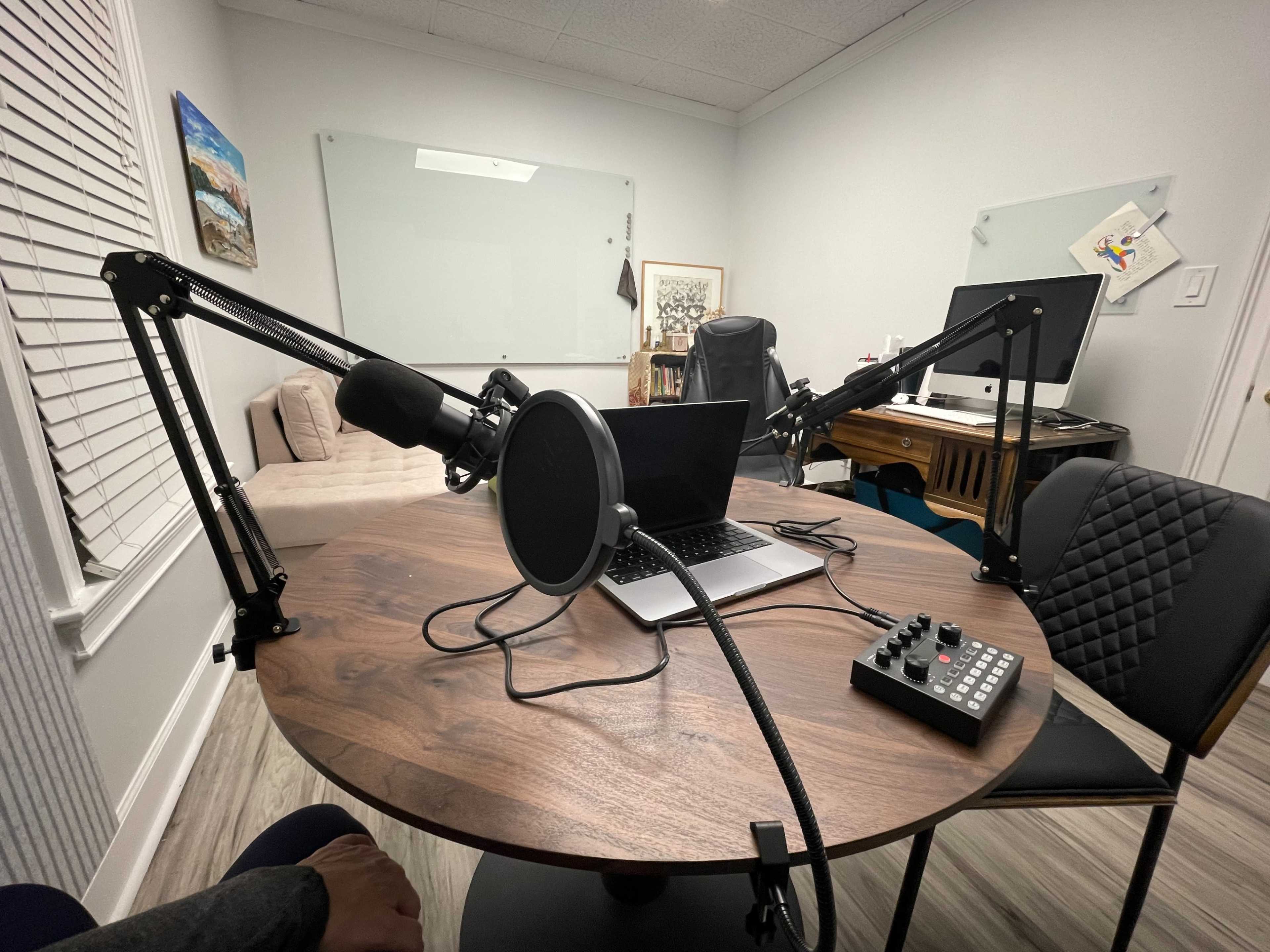 A table with a laptop, microphone, and audio interface is set up in a modern office space featuring a couch and two computer monitors.