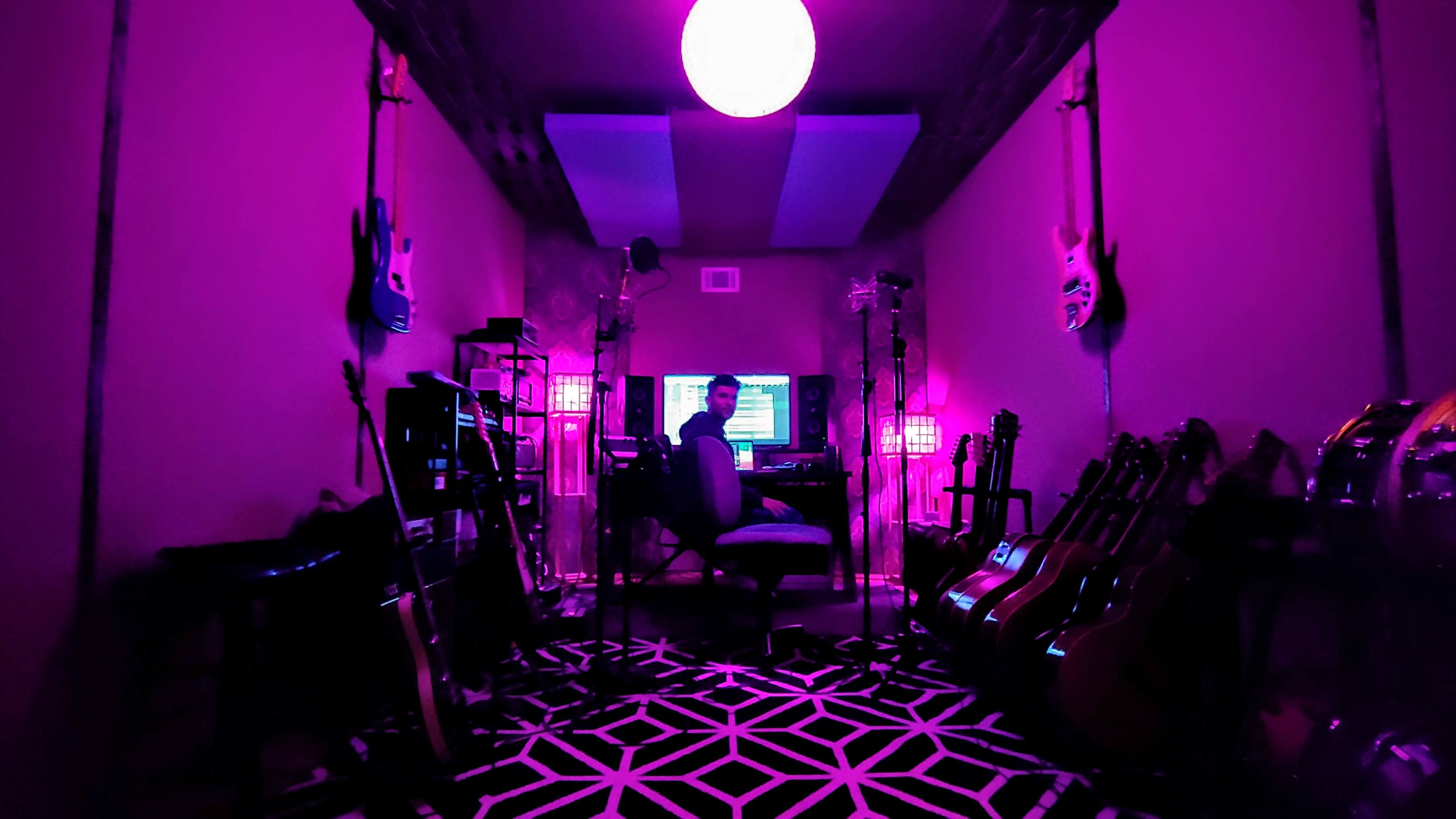 A person sits at a desk in a room filled with musical instruments, illuminated by purple lighting and featuring a geometric patterned floor.