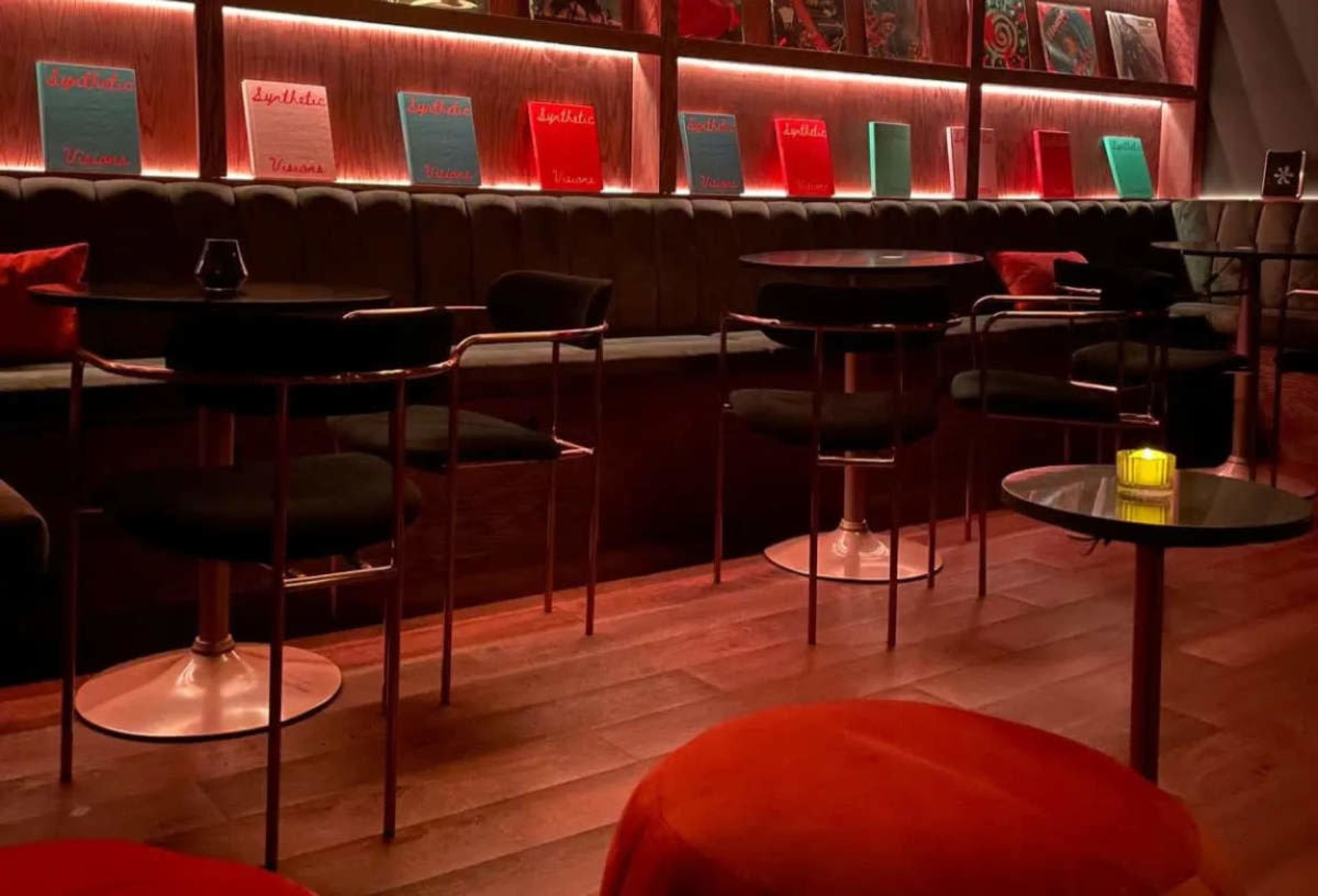 The image shows a dimly lit lounge area with black tables and red upholstered seating, featuring illuminated shelves lined with colorful menus or records.