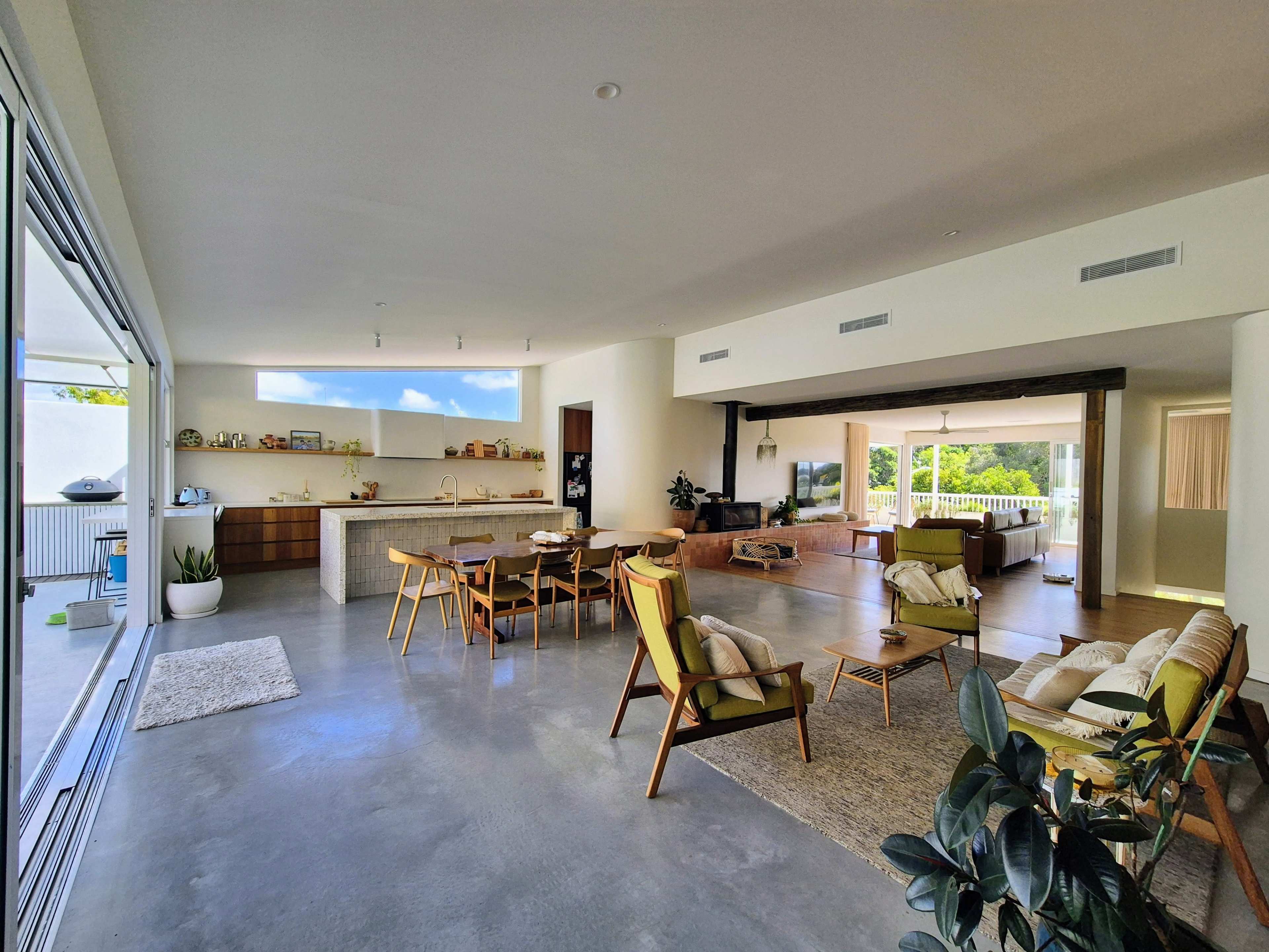 The image shows a spacious living area with a modern kitchen, dining table, and various seating arrangements, featuring large windows that allow natural light to enter.
