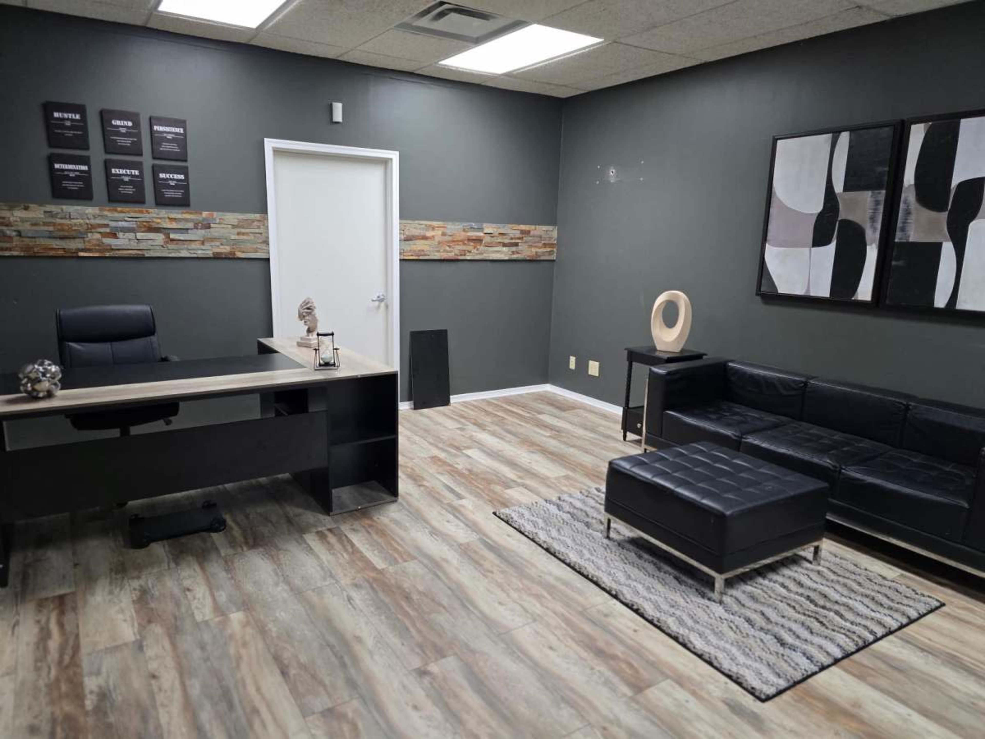 The image shows a modern office interior featuring a black desk with a chair, a black leather sofa, and a textured rug on a wooden floor.