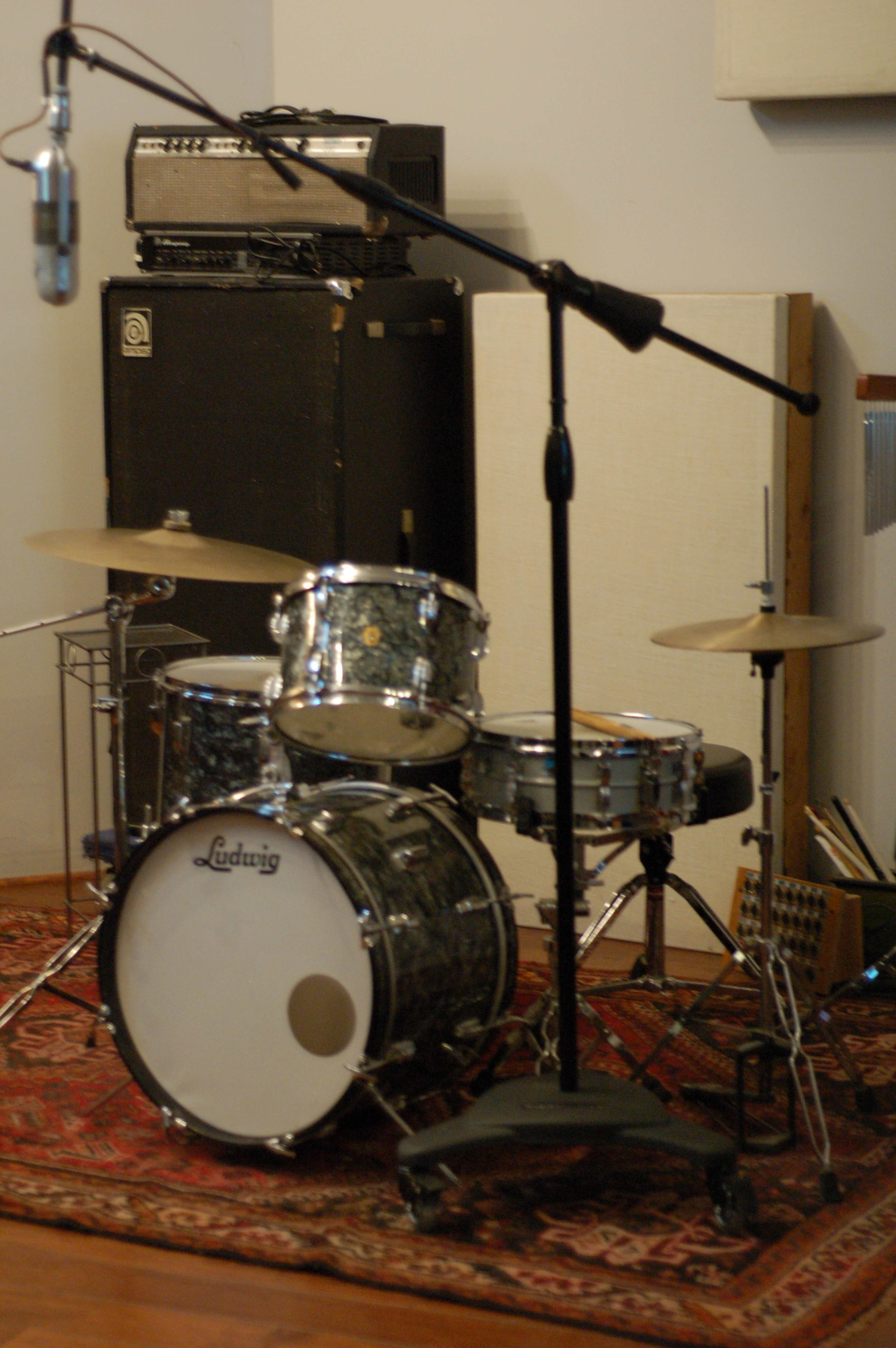 A Ludwig drum set is positioned in front of a microphone with amplifiers and sound equipment in the background on a patterned rug.