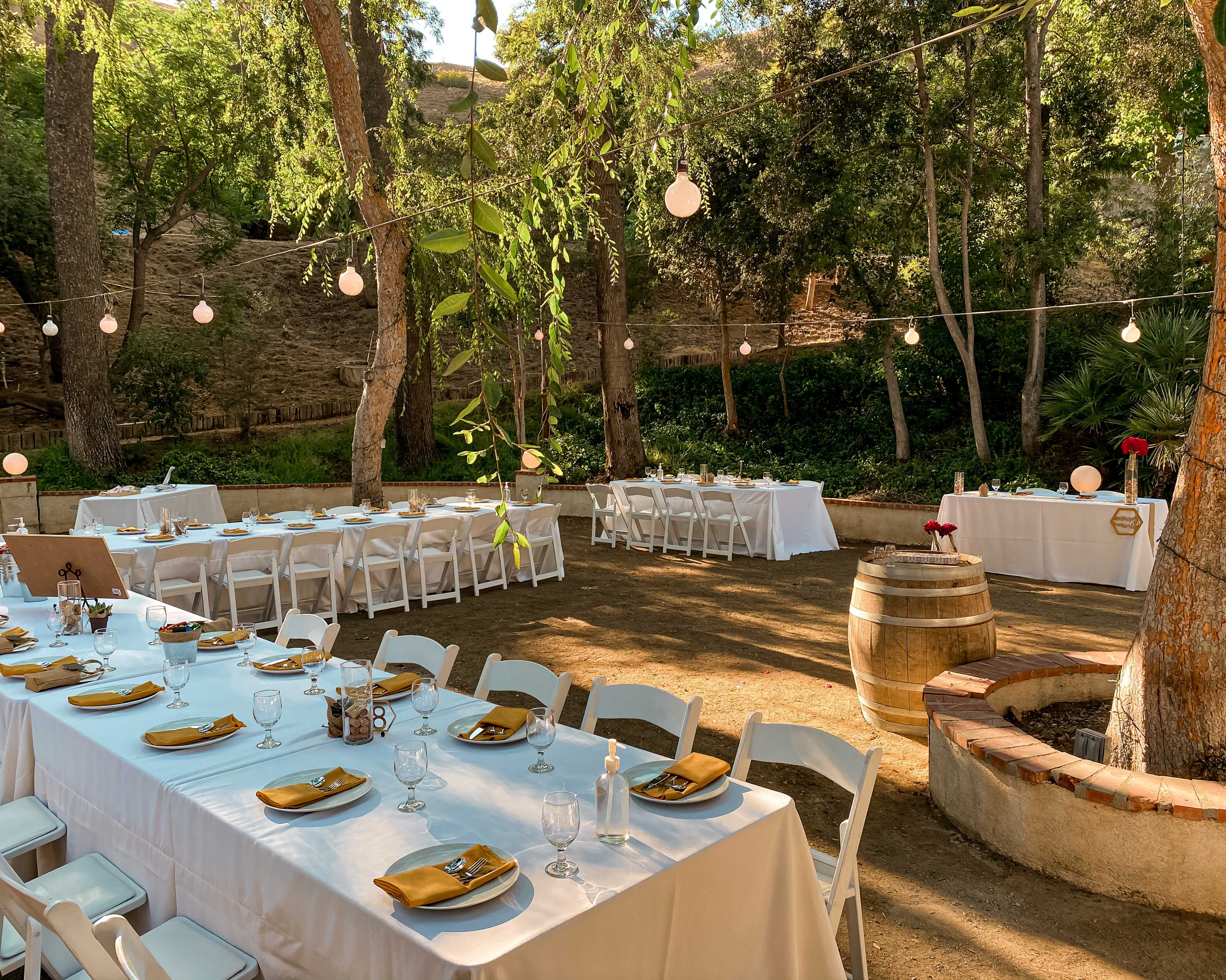A rustic outdoor setting features tables set for dining under string lights, surrounded by trees and lush greenery.