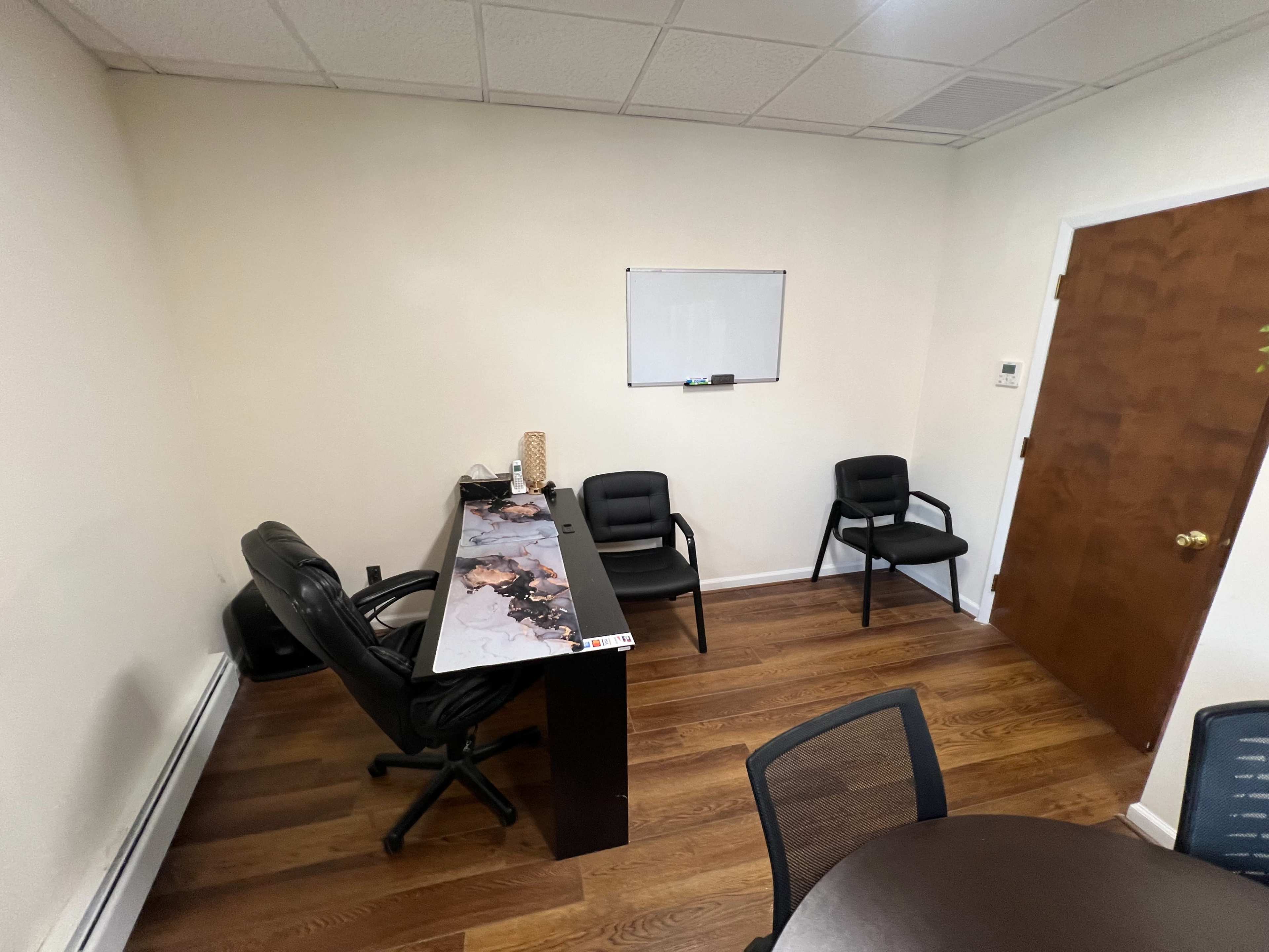 A small office space with a desk, a swivel chair, three black chairs, a whiteboard, and a wooden door.