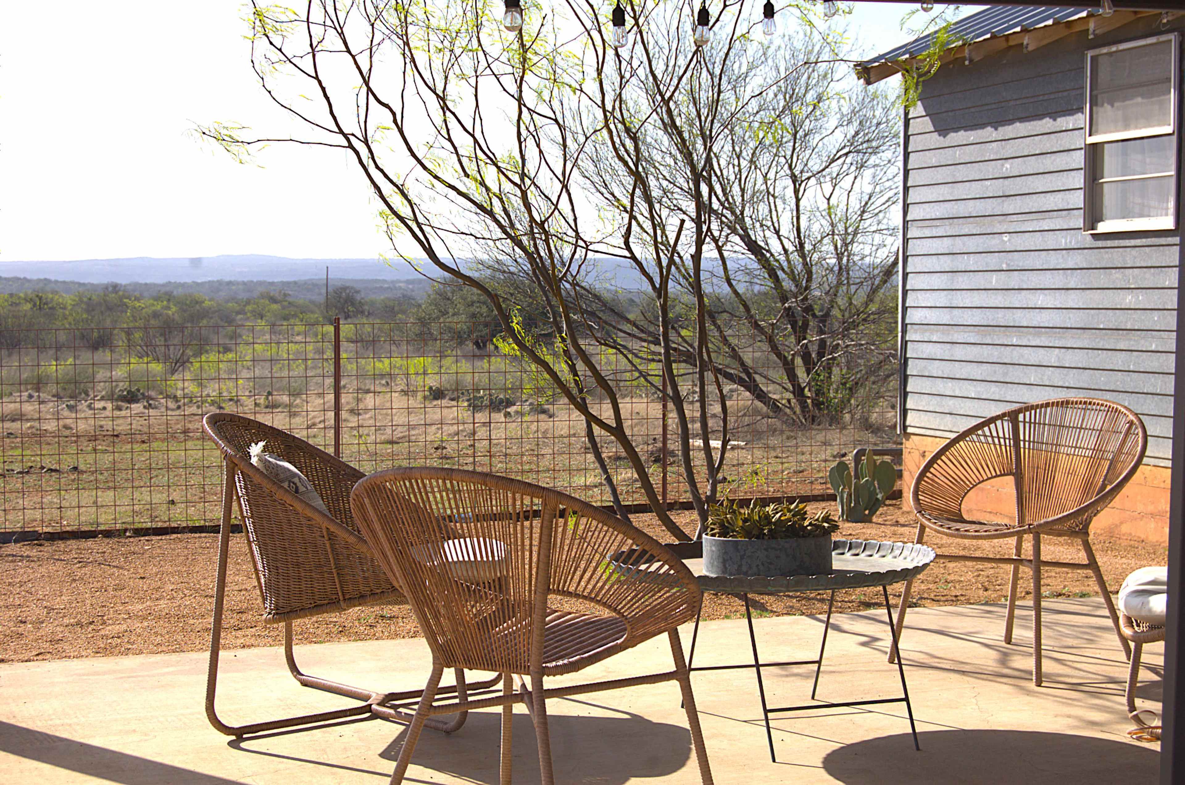 A patio area features three wicker chairs and a small table, facing an open landscape with minimal vegetation and distant hills.