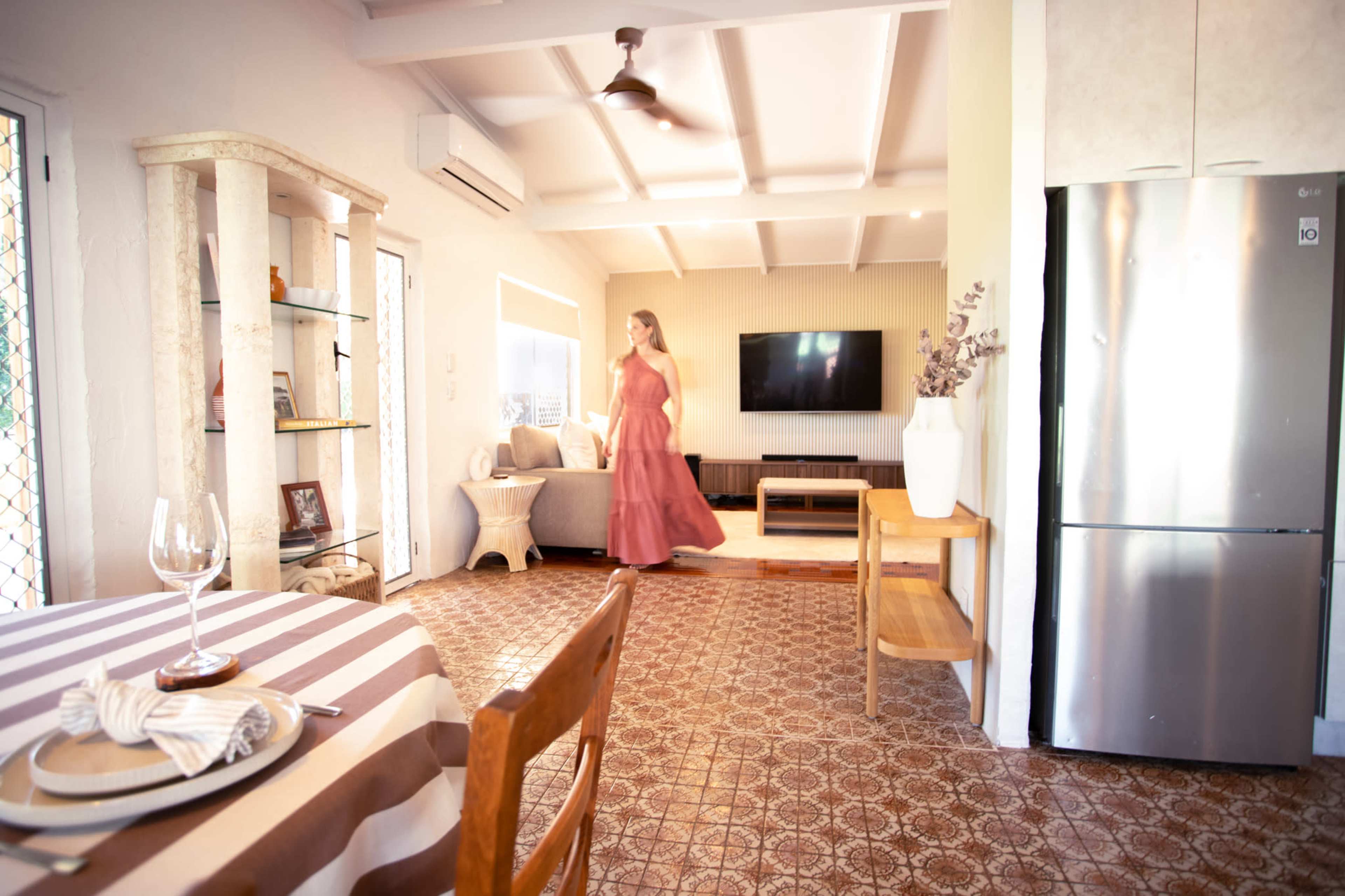 A woman in a long dress walks through a brightly lit living space featuring a gray sofa, a wooden table, and a stainless steel refrigerator.