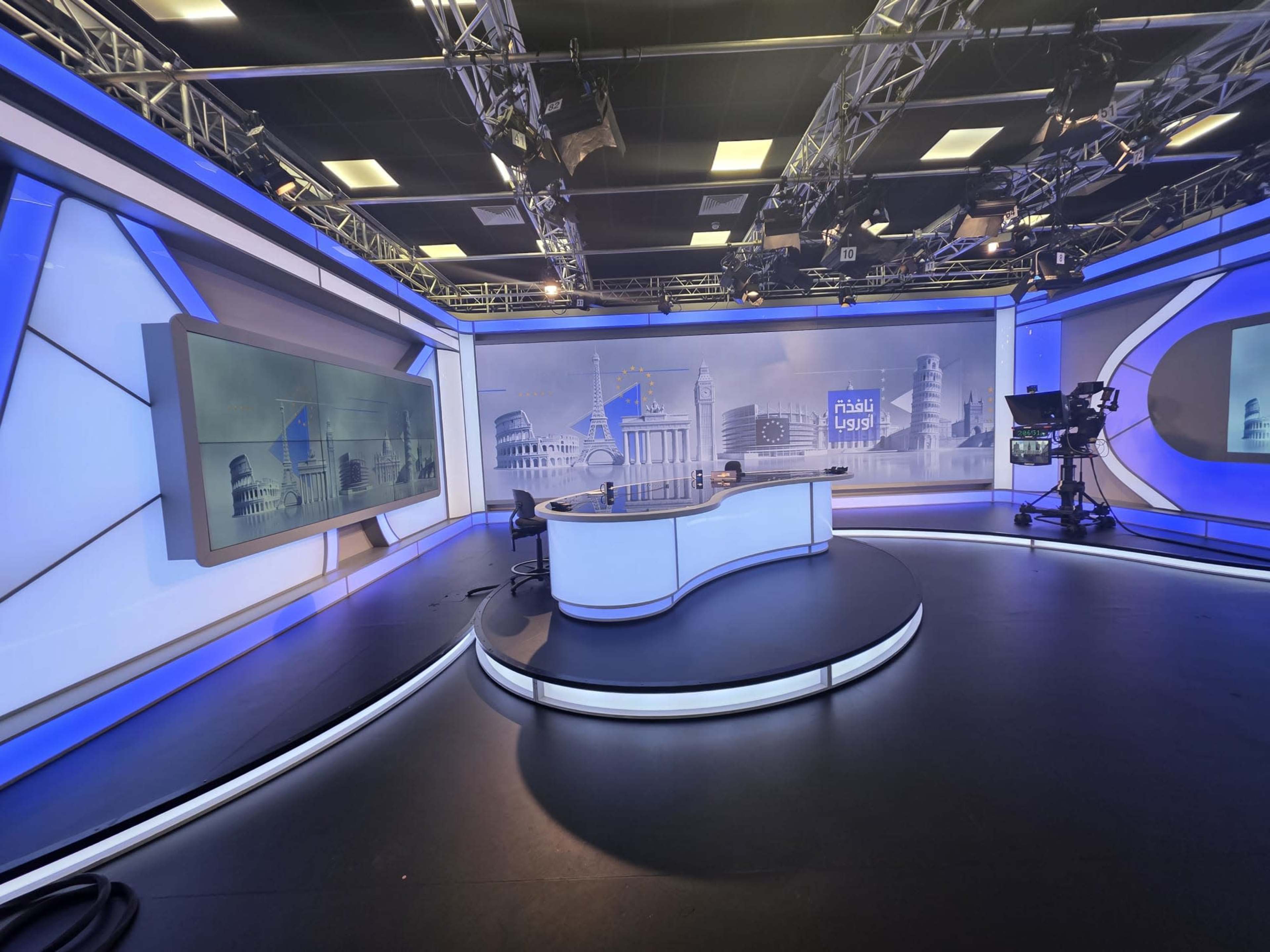The image shows a modern television news studio with a central news desk and multiple screens in a brightly lit environment.
