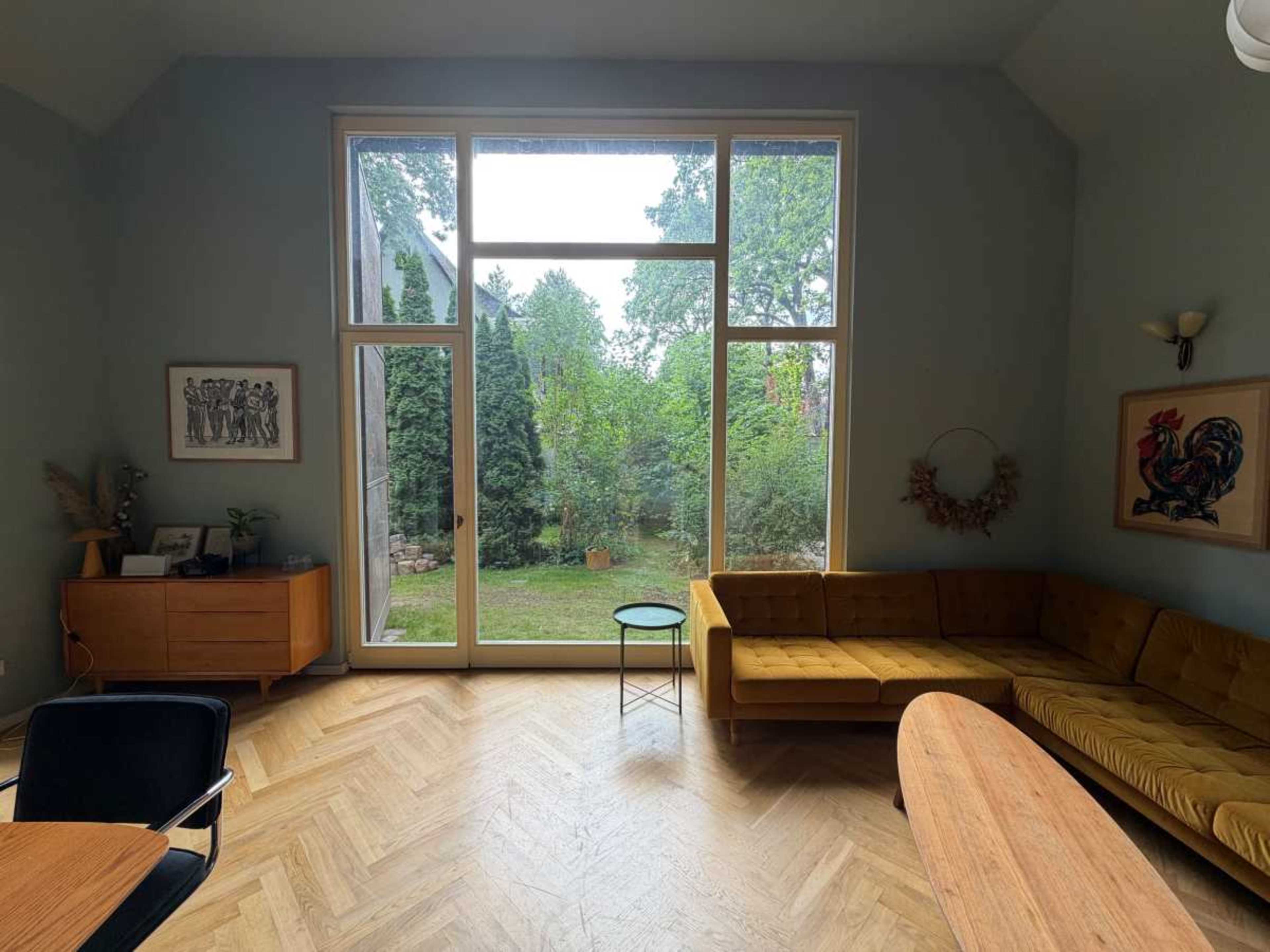 A spacious living room features a large window overlooking a green outdoor space, with a yellow sofa and wooden furniture.