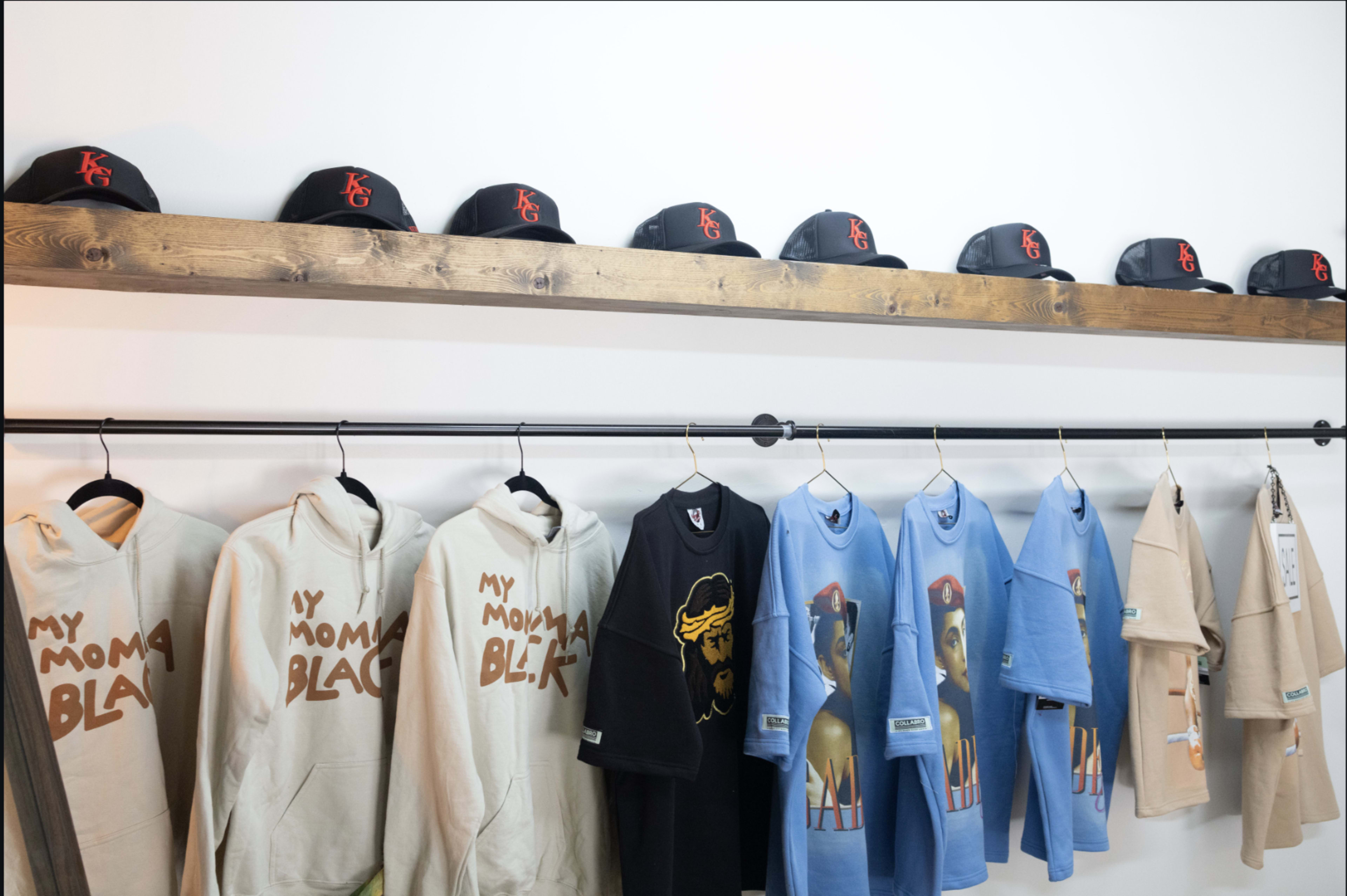 A wooden shelf displays black caps above a rack of hoodies and t-shirts featuring various designs and text.