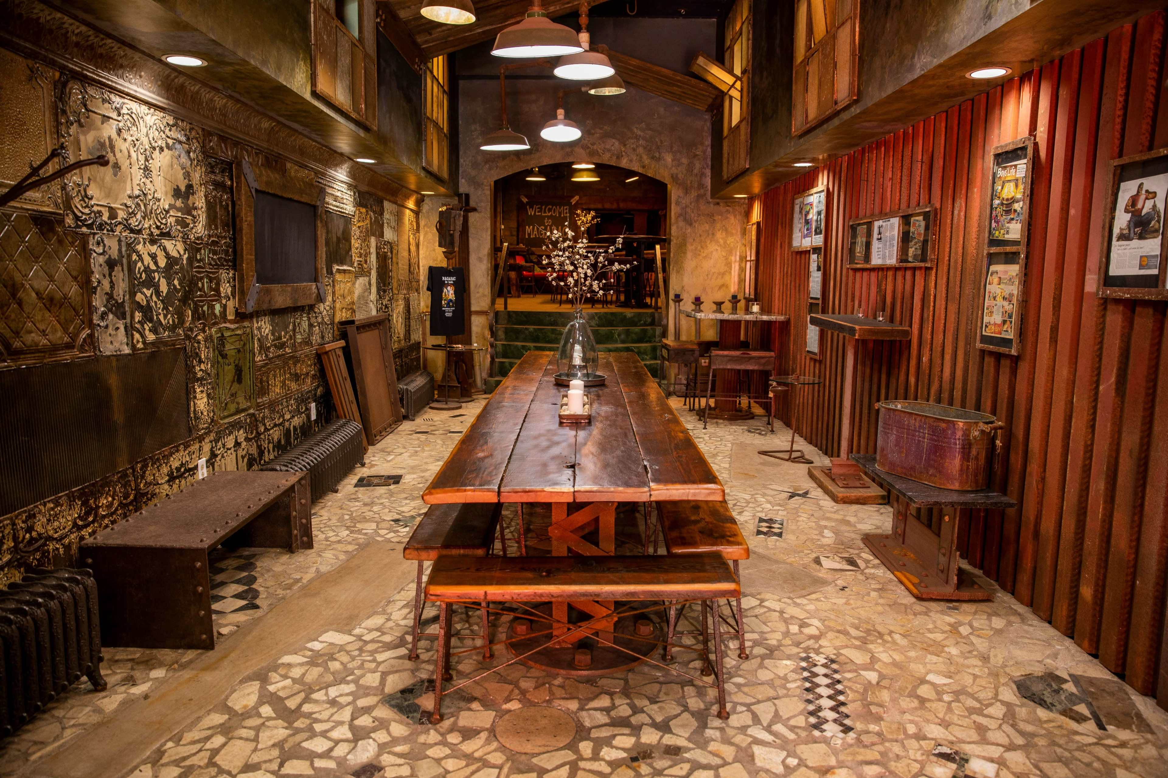 A rustic dining area with a long wooden table, stone flooring, and vintage decor, featuring walls with various textures and framed images.