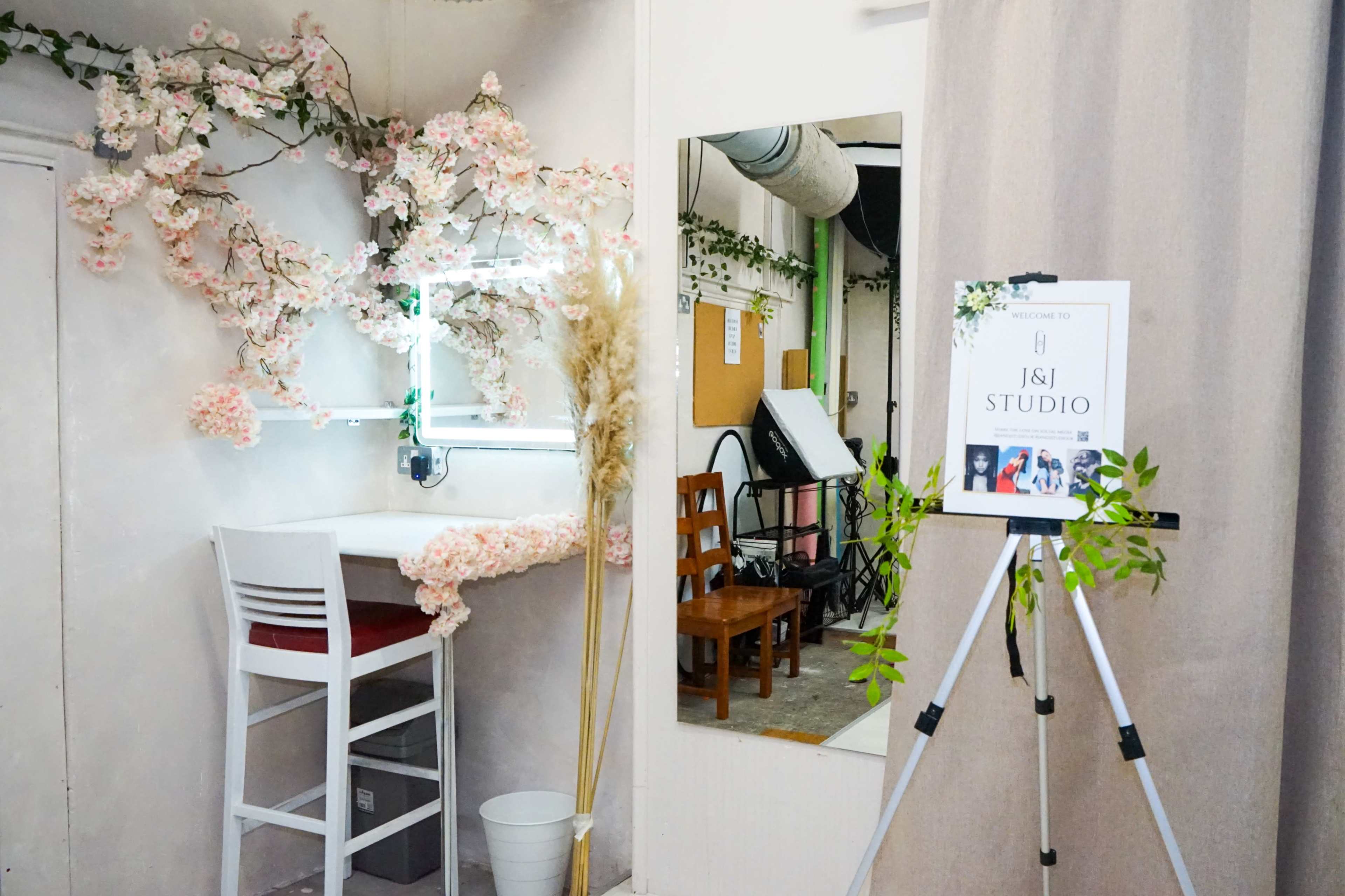 The image shows a studio space featuring a makeup station with a lighted mirror, a decorative floral arrangement, a standing sign that reads "Welcome to J&J Studio," and wooden chairs arranged nearby.
