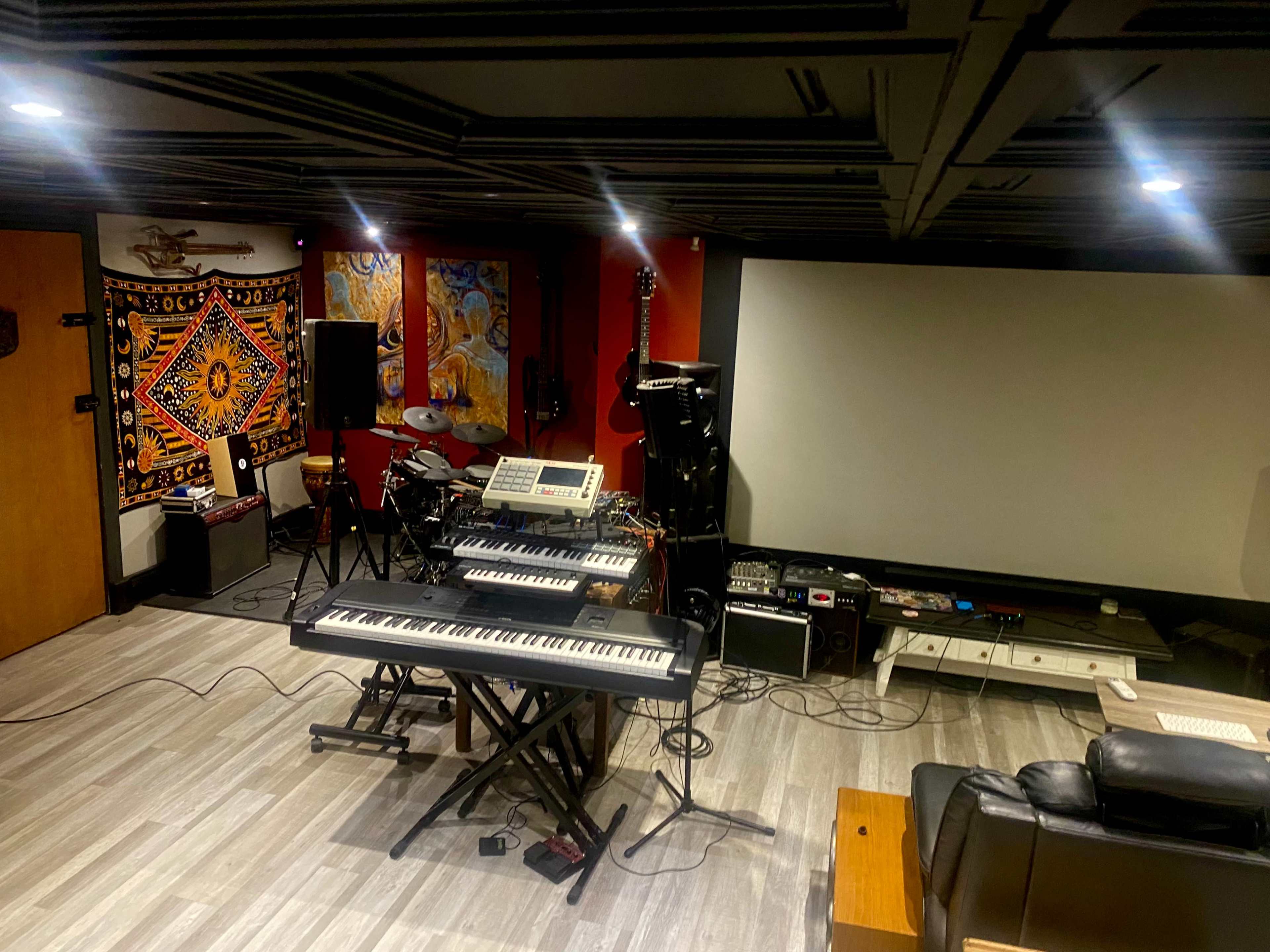 A music studio features a keyboard, electronic drum set, guitar amps, and a large screen in a dimly lit room with a colorful tapestry on one wall.