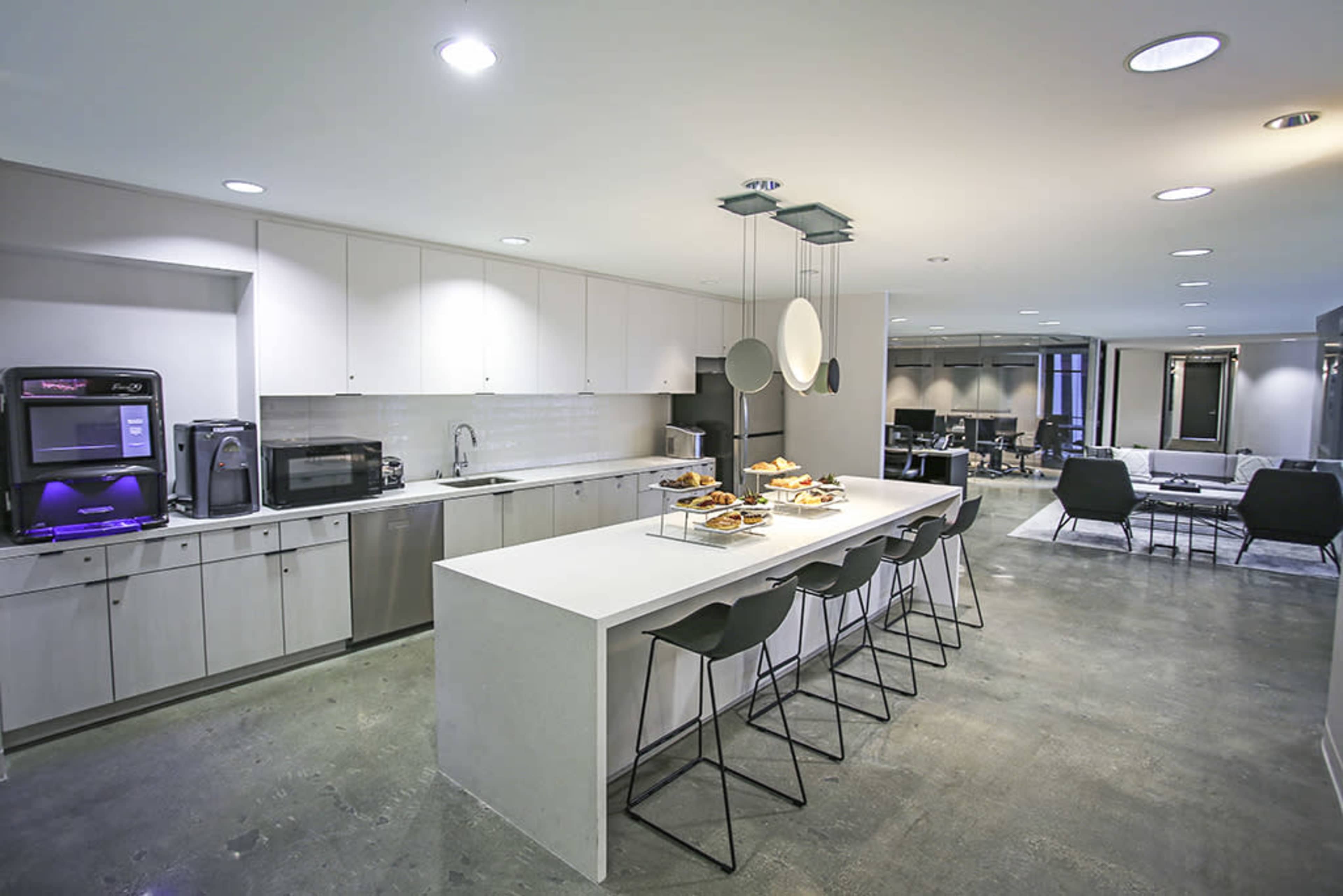 A modern kitchen featuring a long white countertop with bar stools, stainless steel appliances, and a seating area in the background.