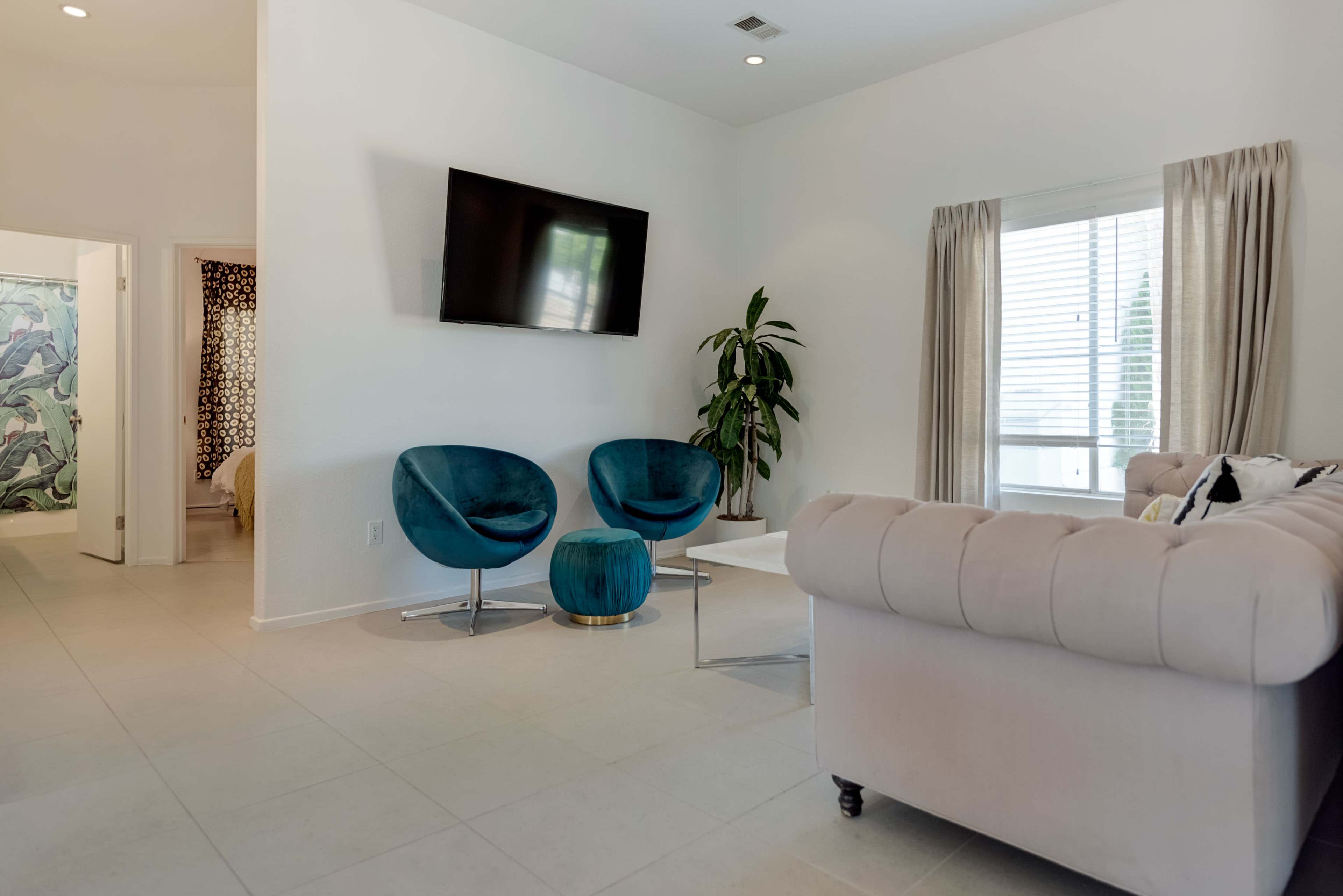 A living room features two teal armchairs, a beige sofa, a mounted television, and a potted plant, with a doorway leading to another room in the background.