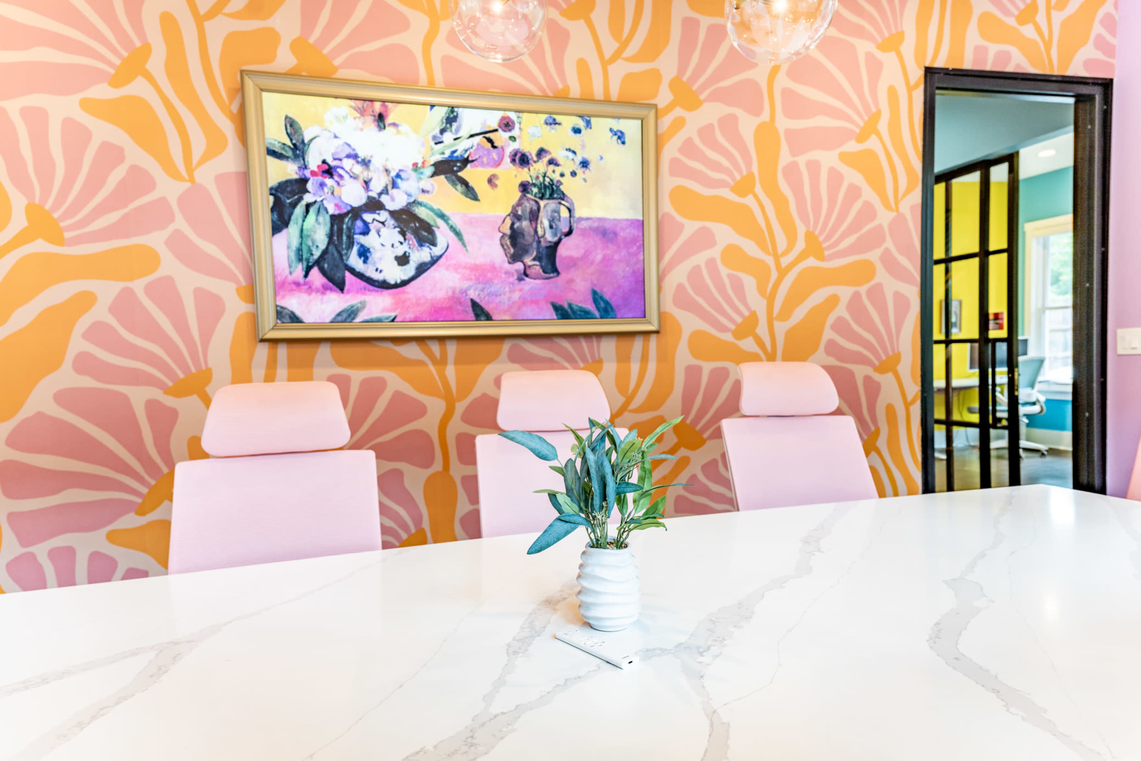 A modern conference room features a marble table, pink chairs, and a colorful abstract painting on the wall, set against a vibrant, patterned backdrop.