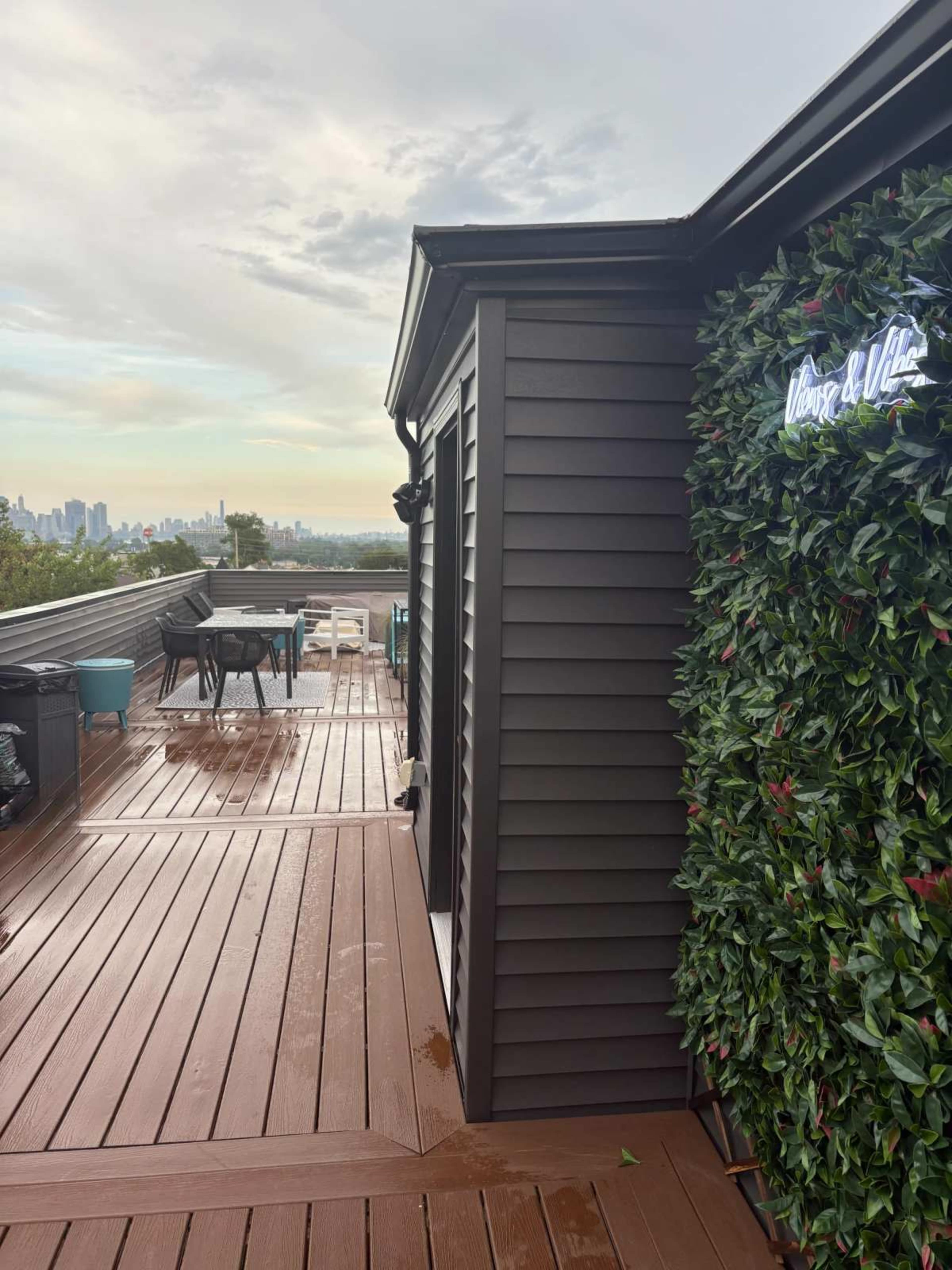 A rooftop deck features wooden flooring, a greenery wall, and views of a city skyline in the distance.