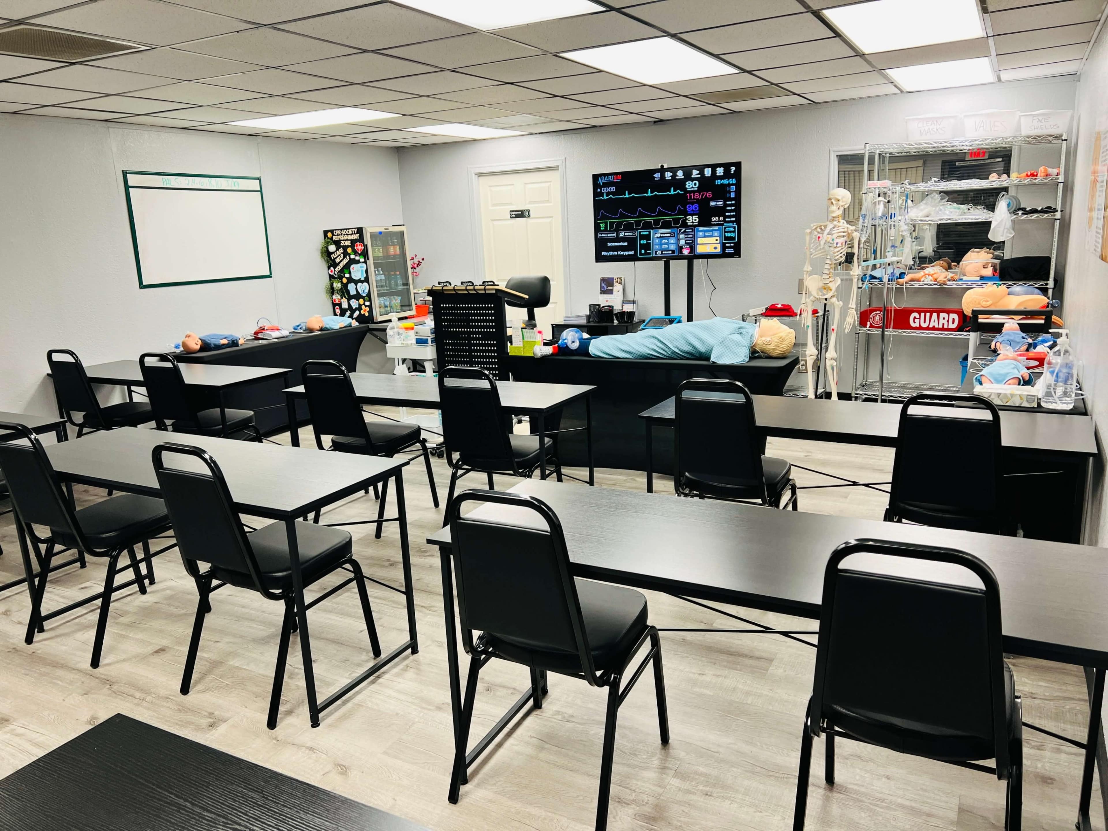 A classroom is arranged with black tables and chairs, featuring medical training supplies and a monitor displaying health data.