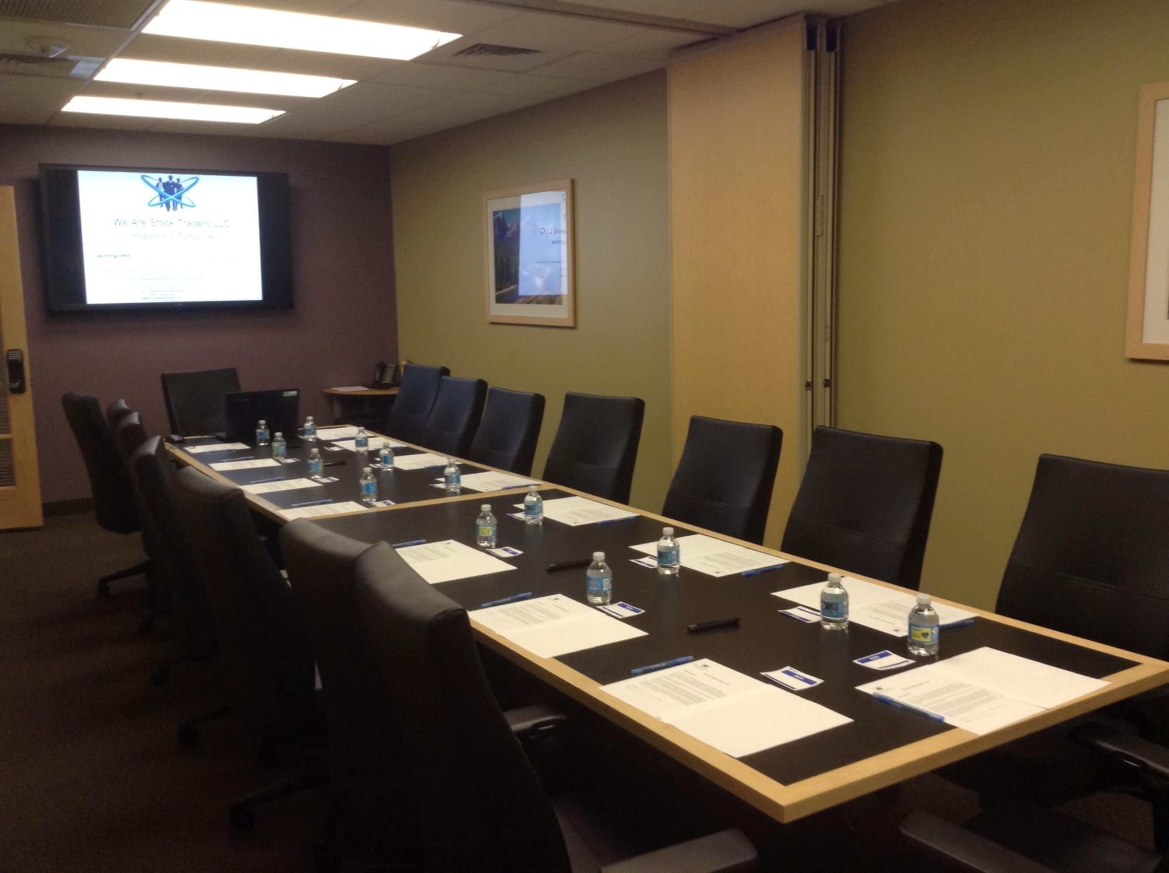 A large conference room features a long table set with water bottles and papers, with a presentation screen displayed at one end.