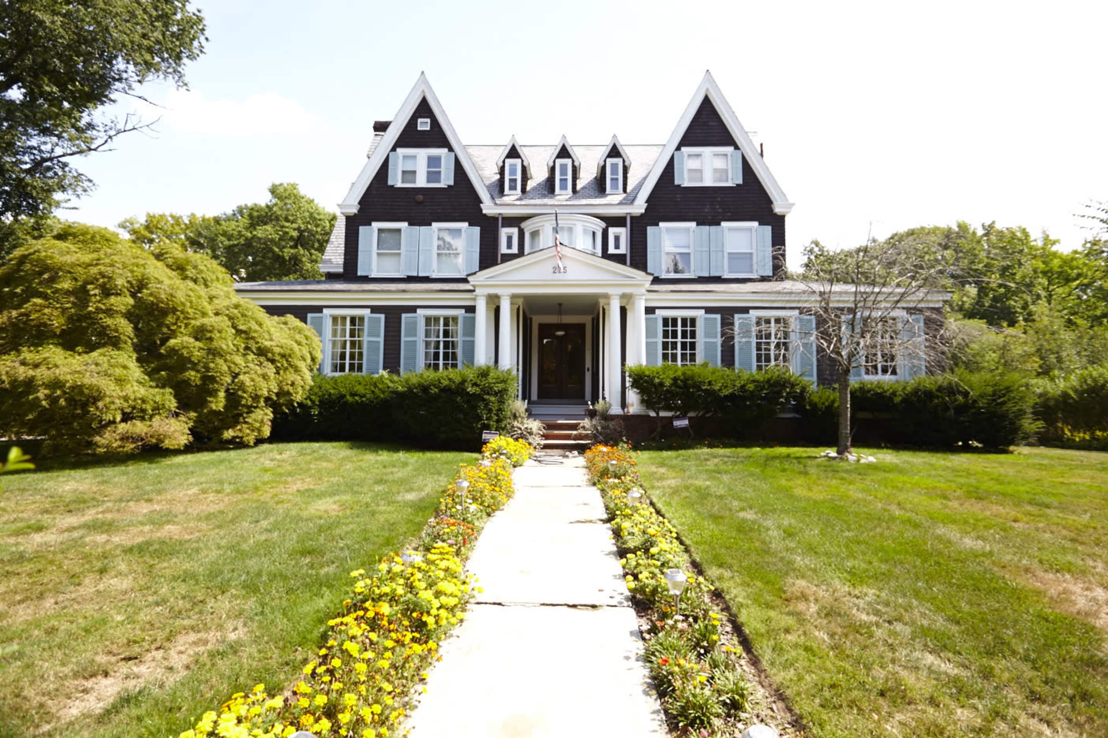 A large, dark-colored house with a steep roof and multiple gables is set behind a well-maintained front yard and pathway lined with colorful flowers.