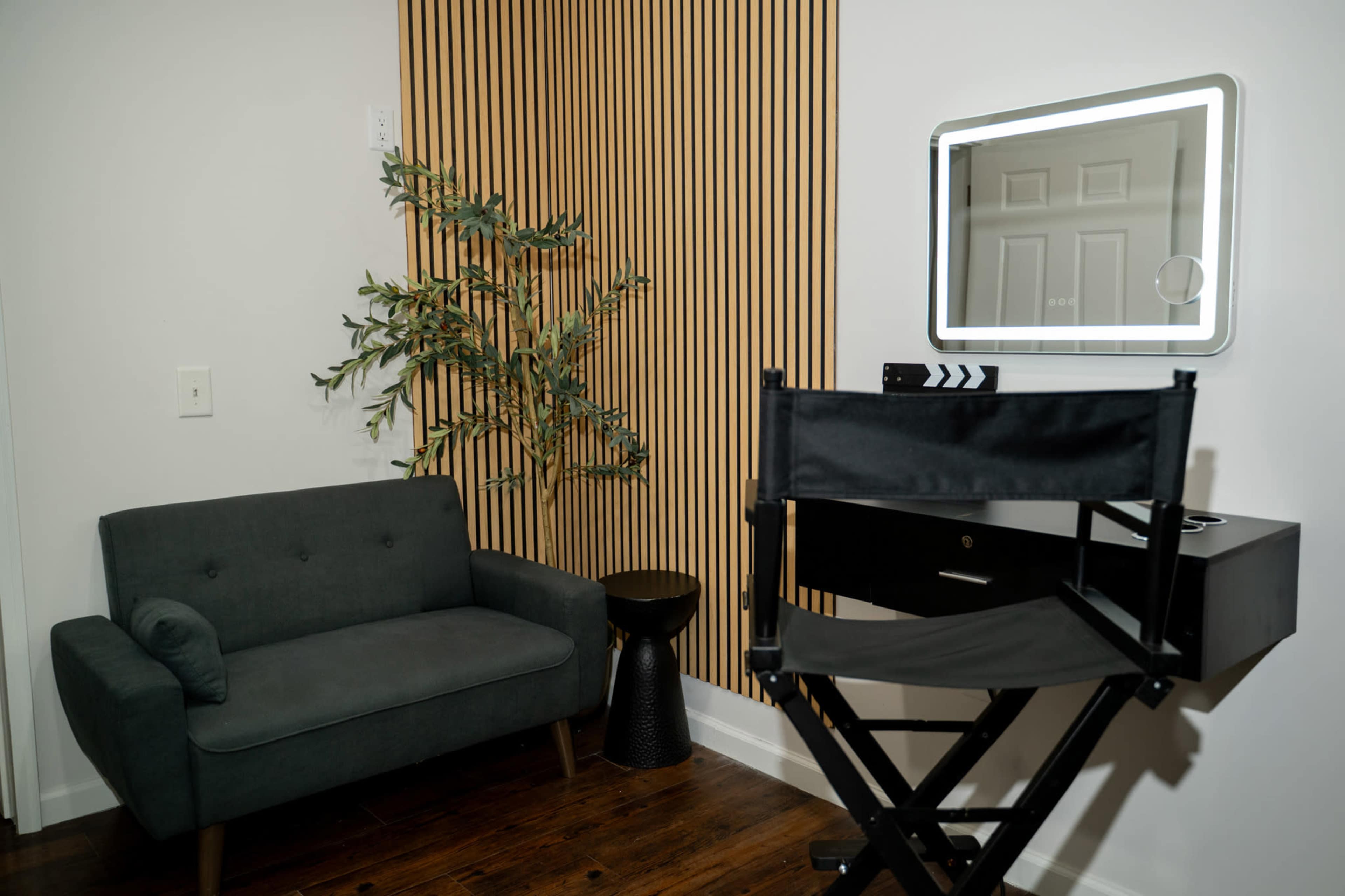 The image shows a small room with a green sofa, a black stool, a director's chair, and a backlit mirror on a wall-mounted desk.