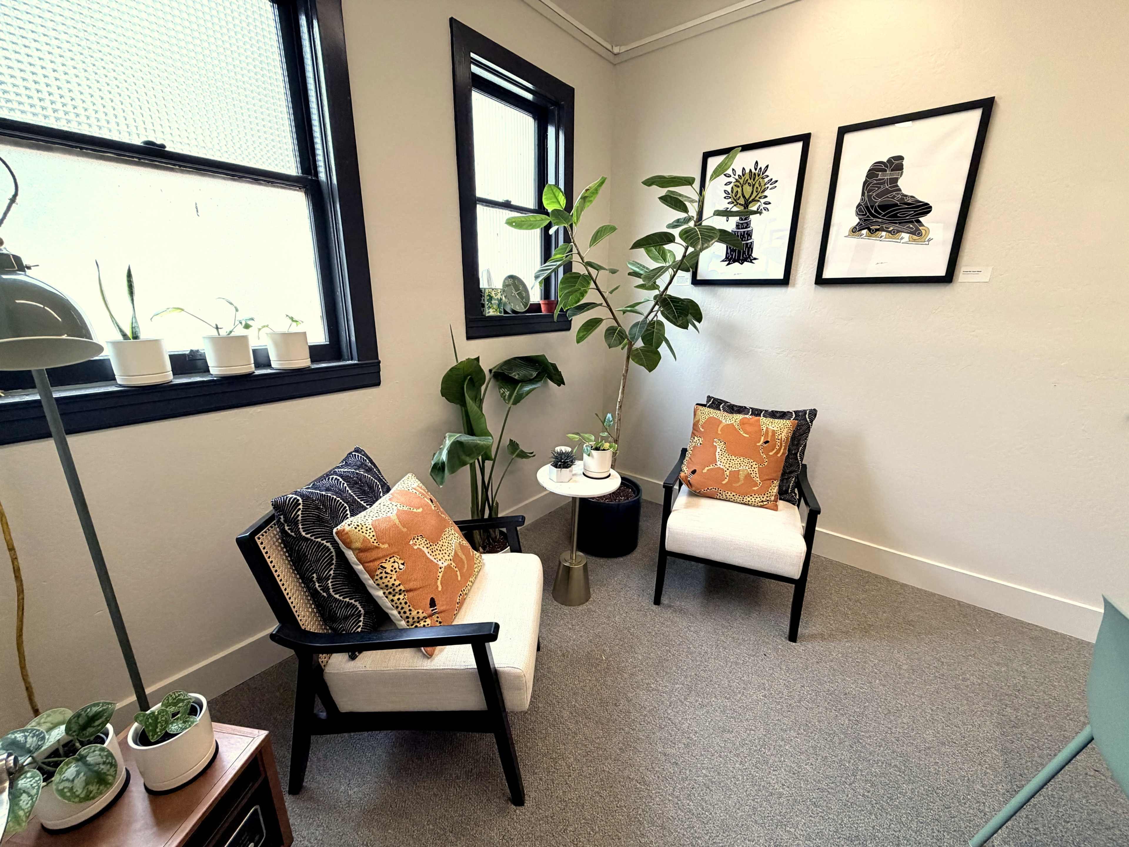 The image shows a cozy corner of a room featuring two chairs with decorative pillows, a small side table, and houseplants, along with framed art on the wall.